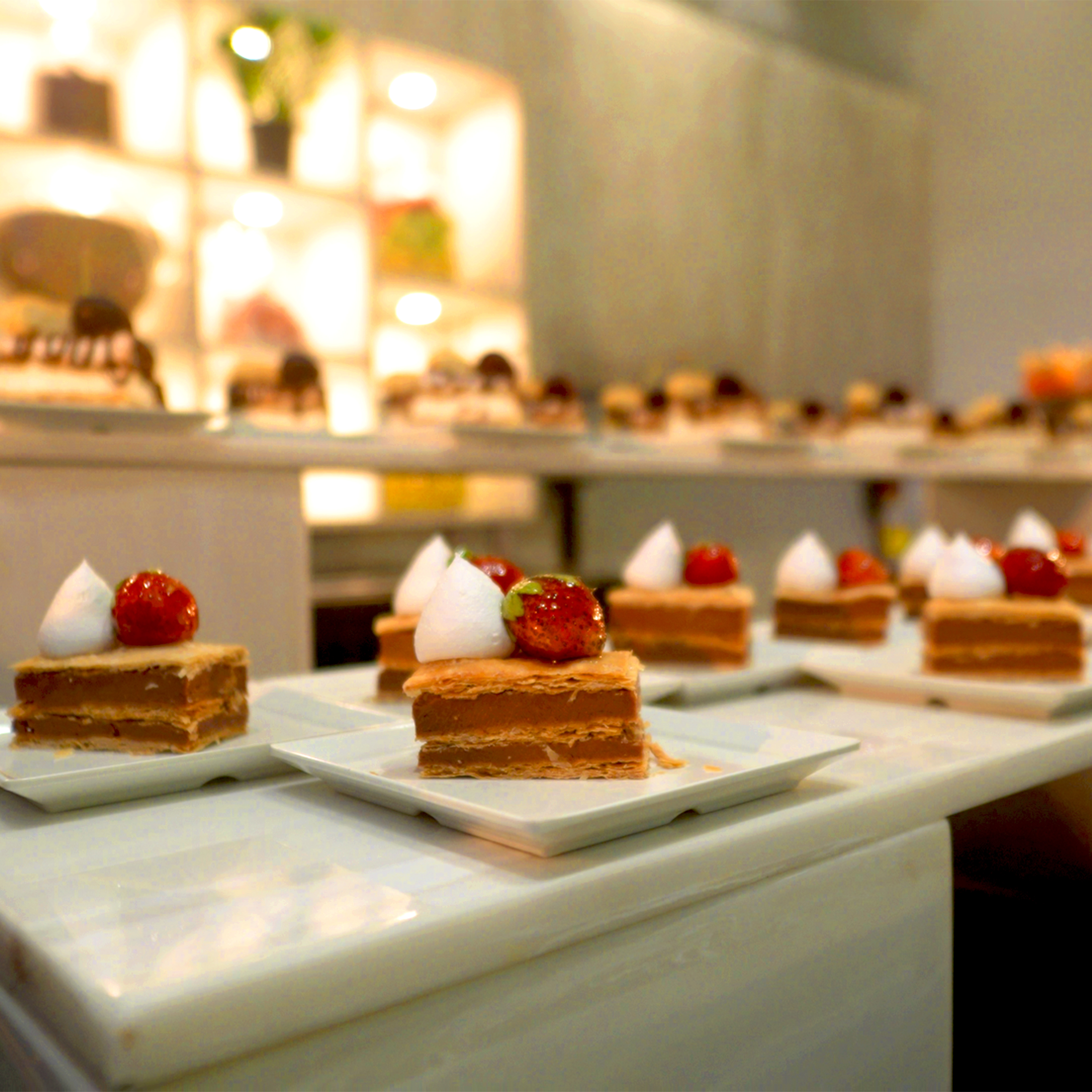 Multiple square slices of layered cake topped with white cream and strawberries on white square plates.