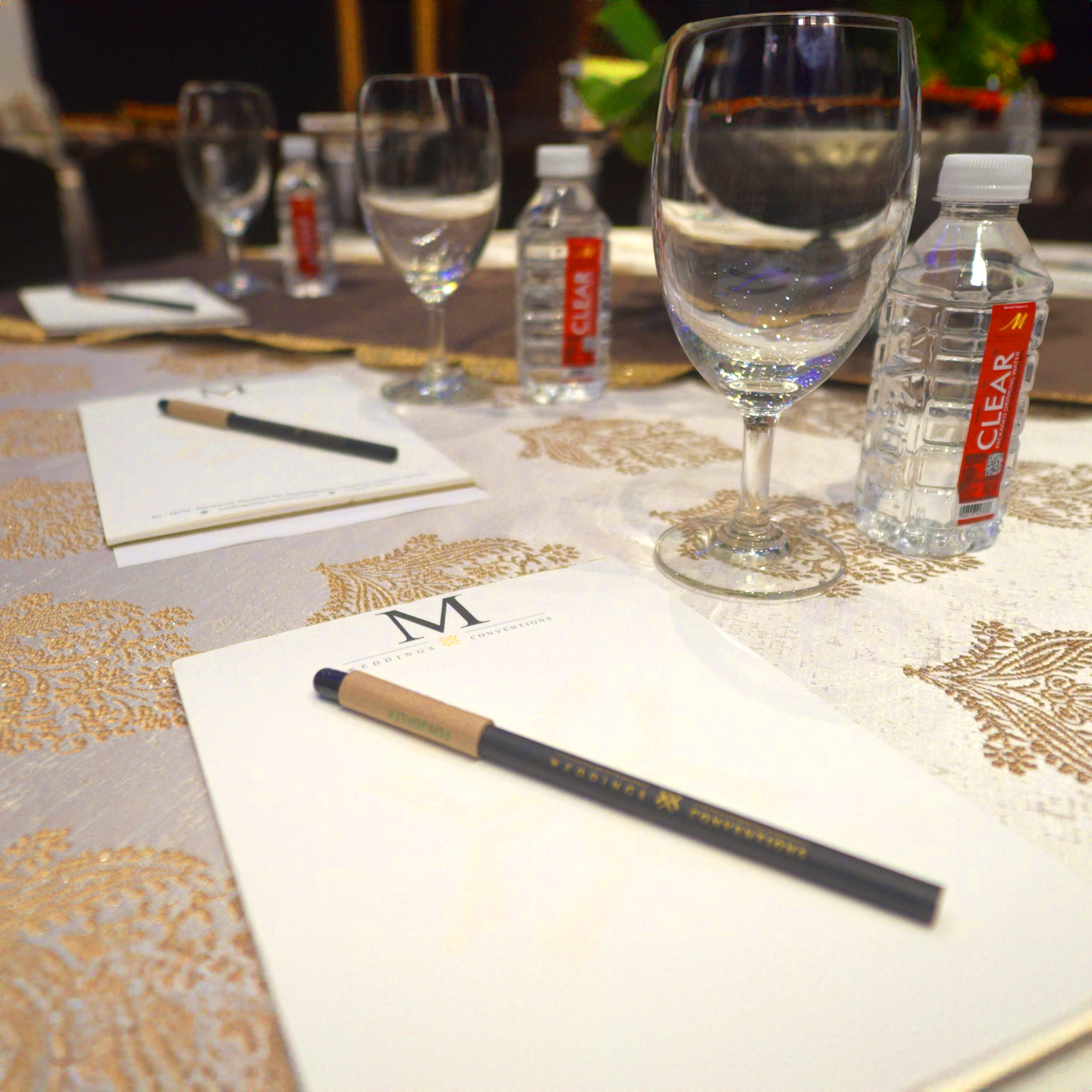 Table set with notebooks and pens labeled 'Weddings & Conventions', empty wine glasses, and bottled water with red labels.