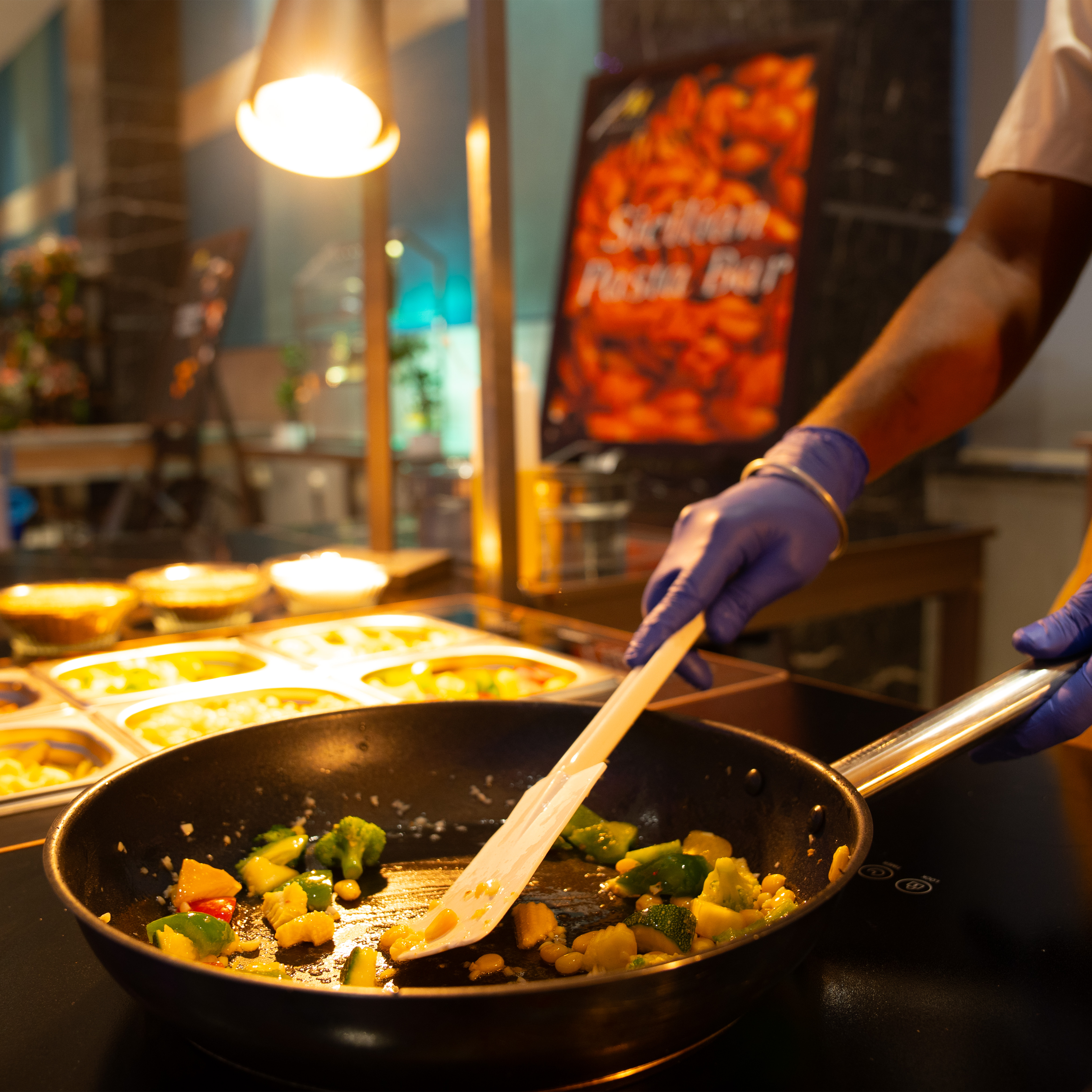 Cook wearing blue gloves stirring mixed vegetables in a black frying pan on a stovetop.