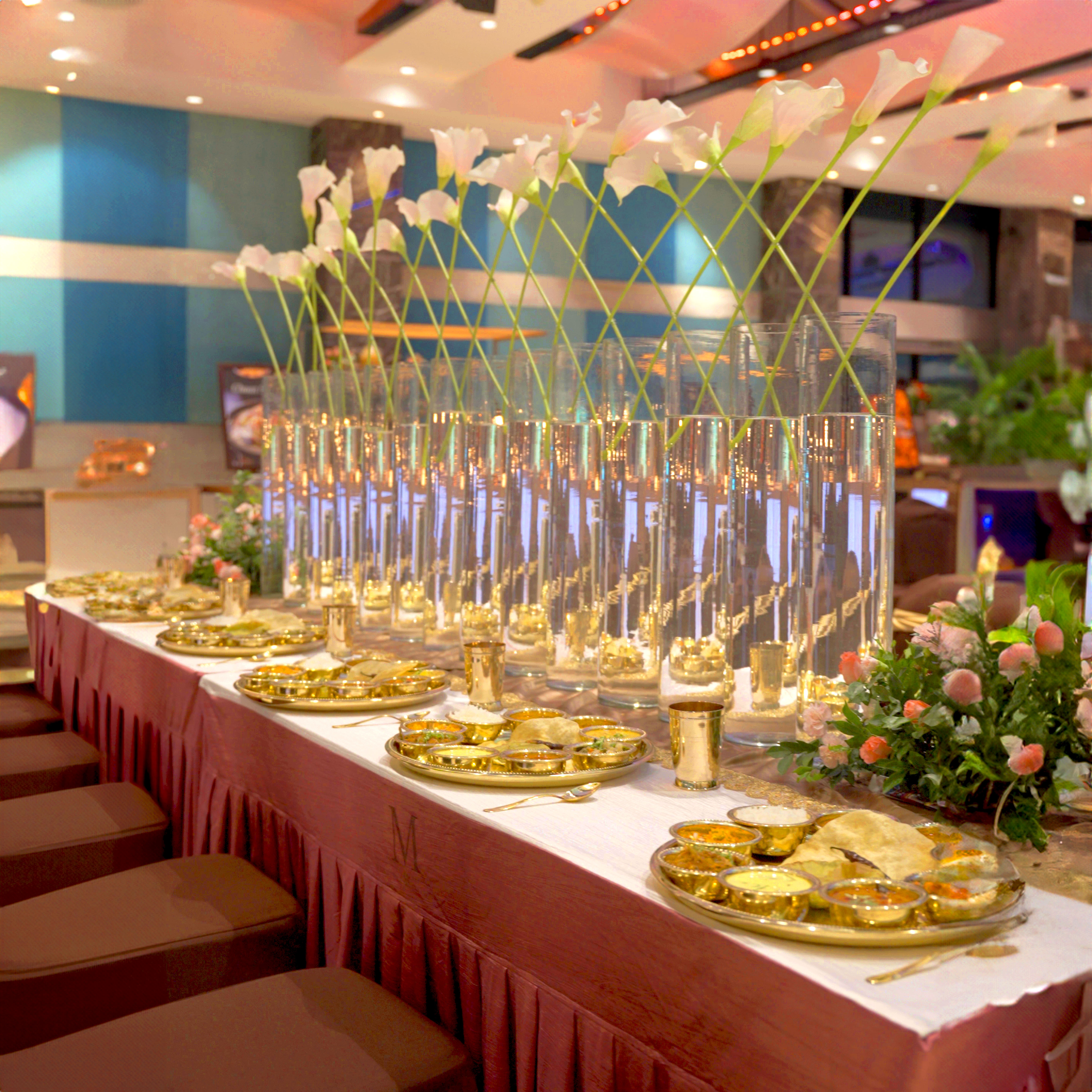 Elegant dining table set with gold plates of Indian food, tall glass vases holding white calla lilies, and floral arrangements in a banquet hall.