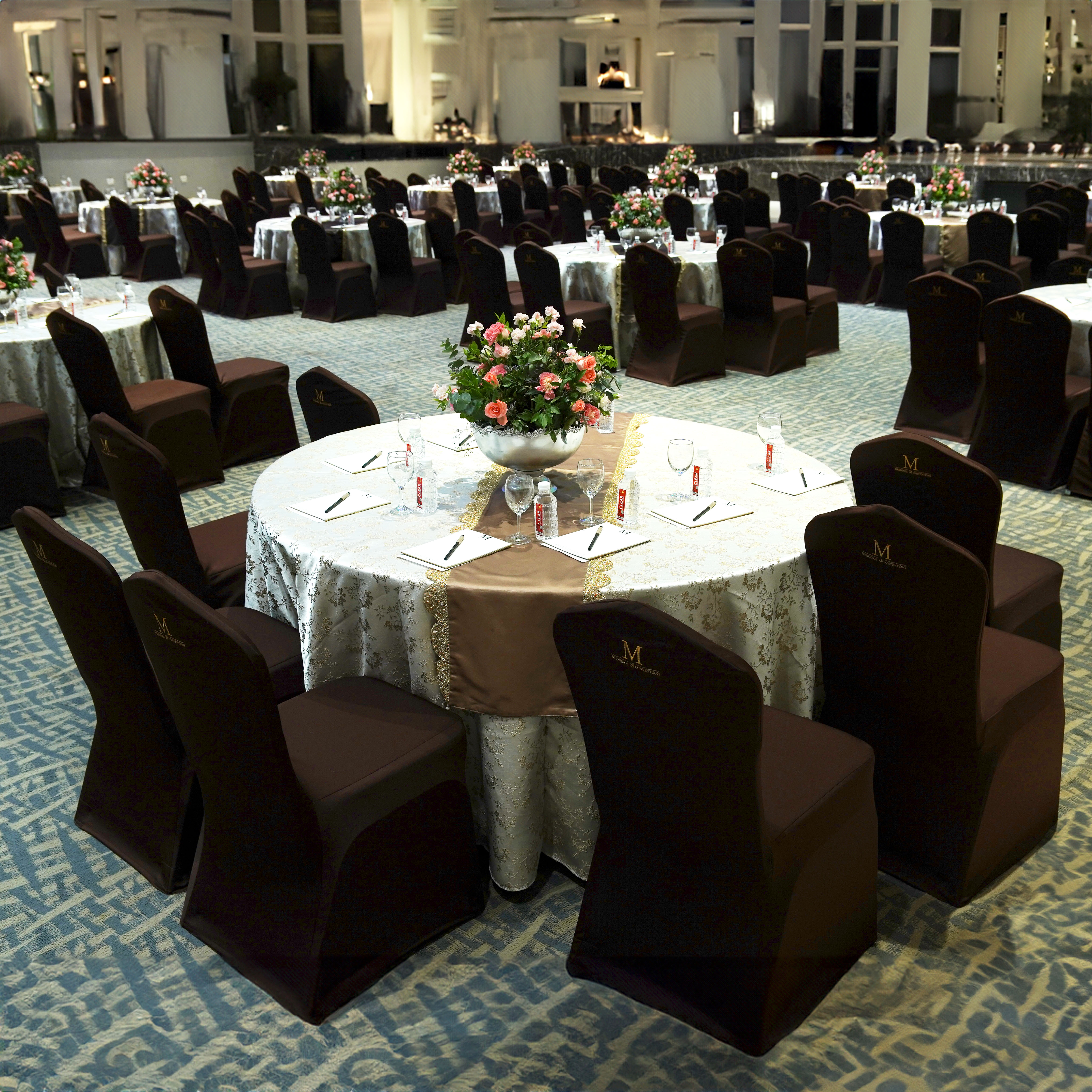 Round banquet table with floral centerpiece, notebooks, pens, water bottles, and covered chairs set in an elegant conference room.
