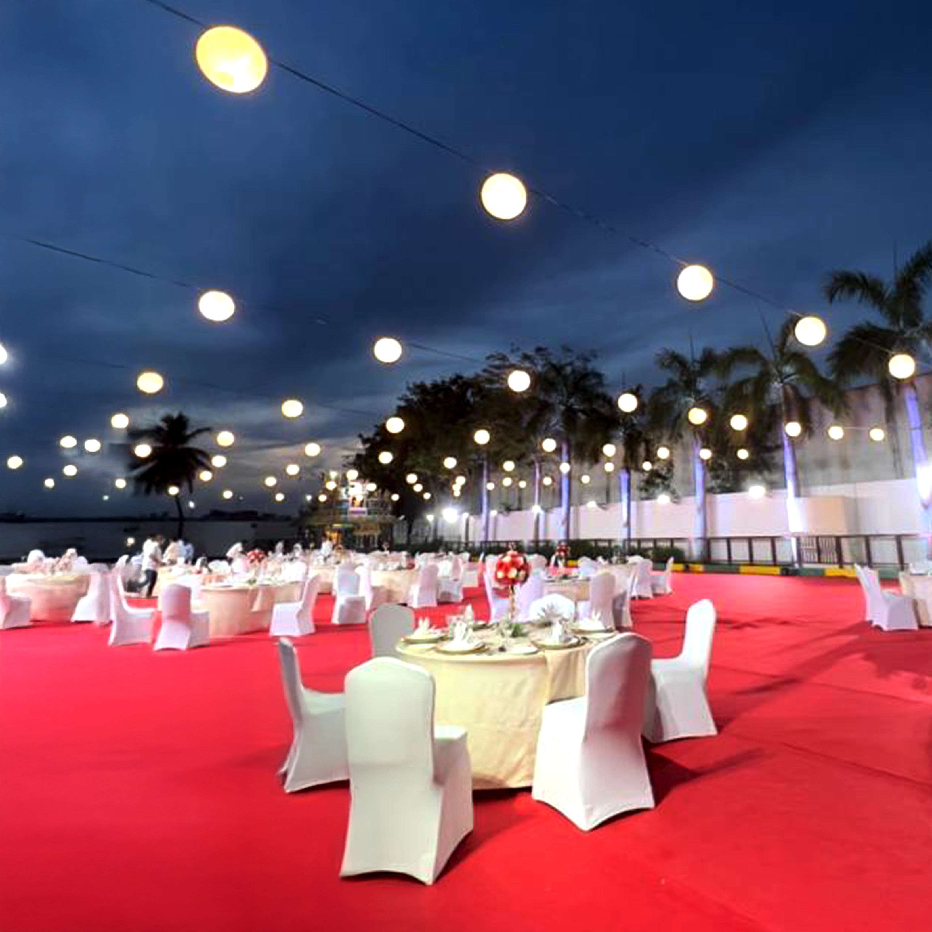 Outdoor evening banquet setup with round tables covered in beige tablecloths and white chairs on a red carpet under hanging string lights and palm trees.