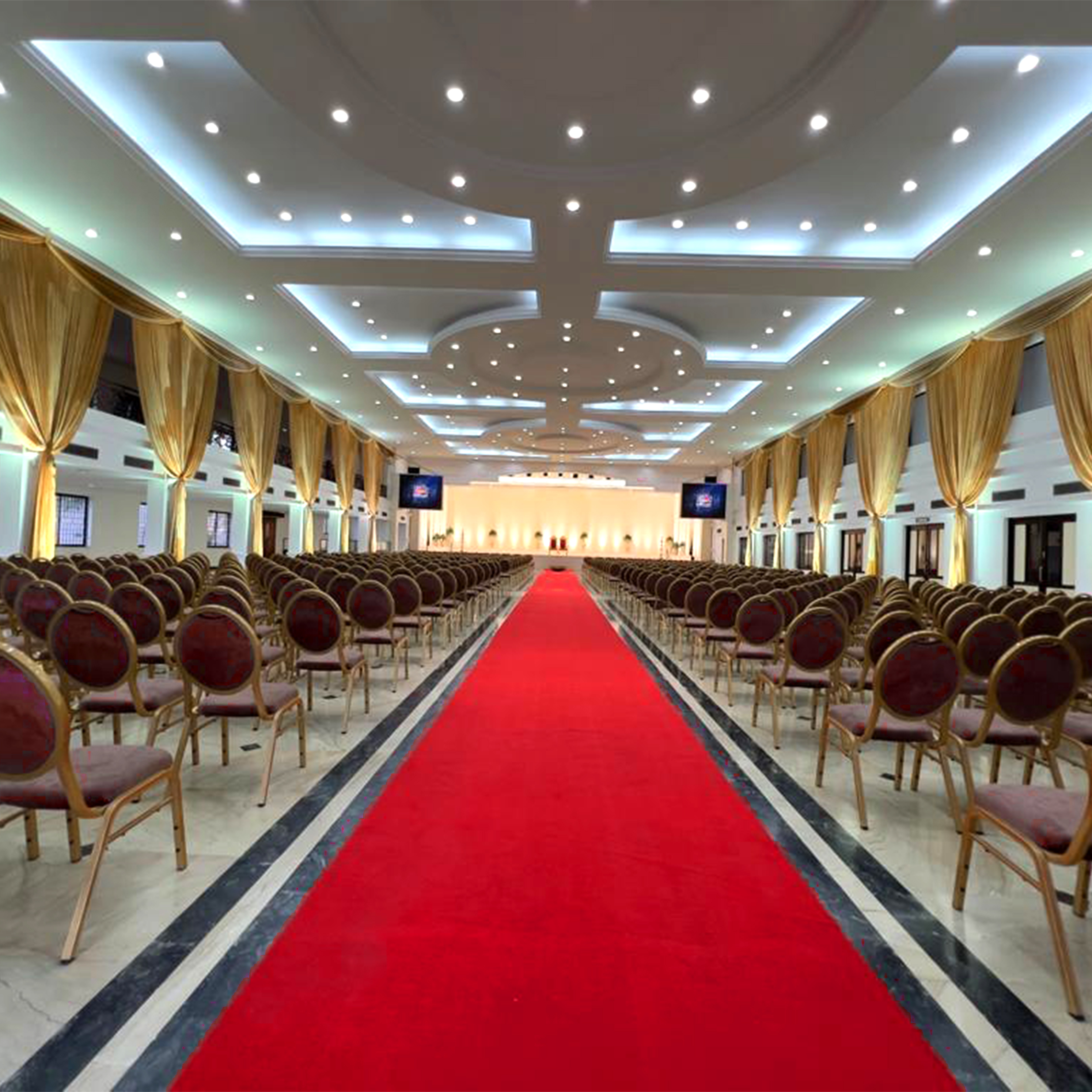 Large elegant auditorium with rows of red and gold chairs facing a stage, illuminated ceiling, and a red carpet aisle.