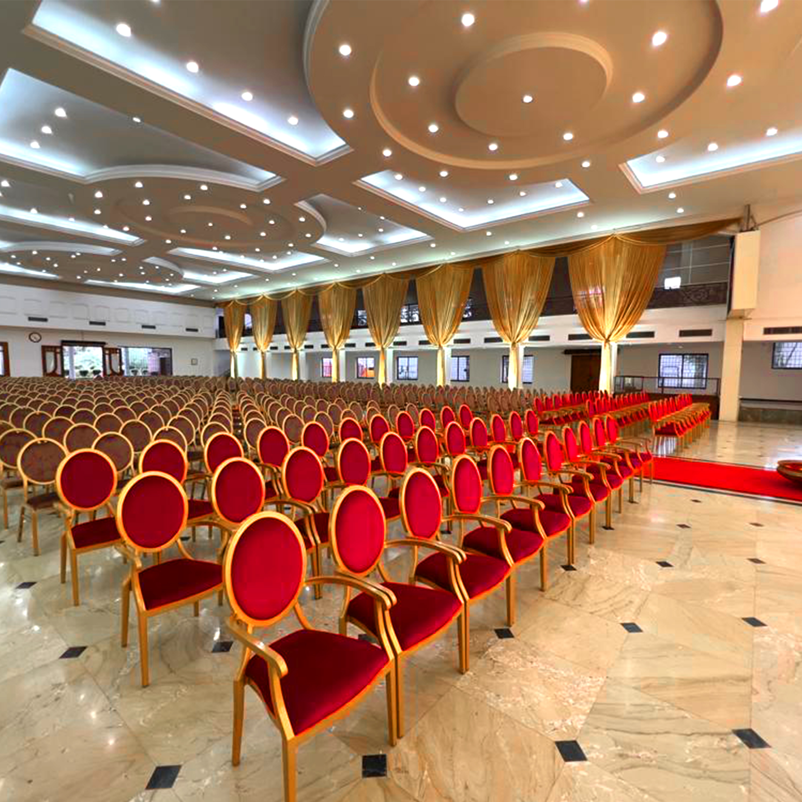 Spacious event hall with rows of red and gold chairs on polished marble floor under a ceiling with multiple recessed lights.