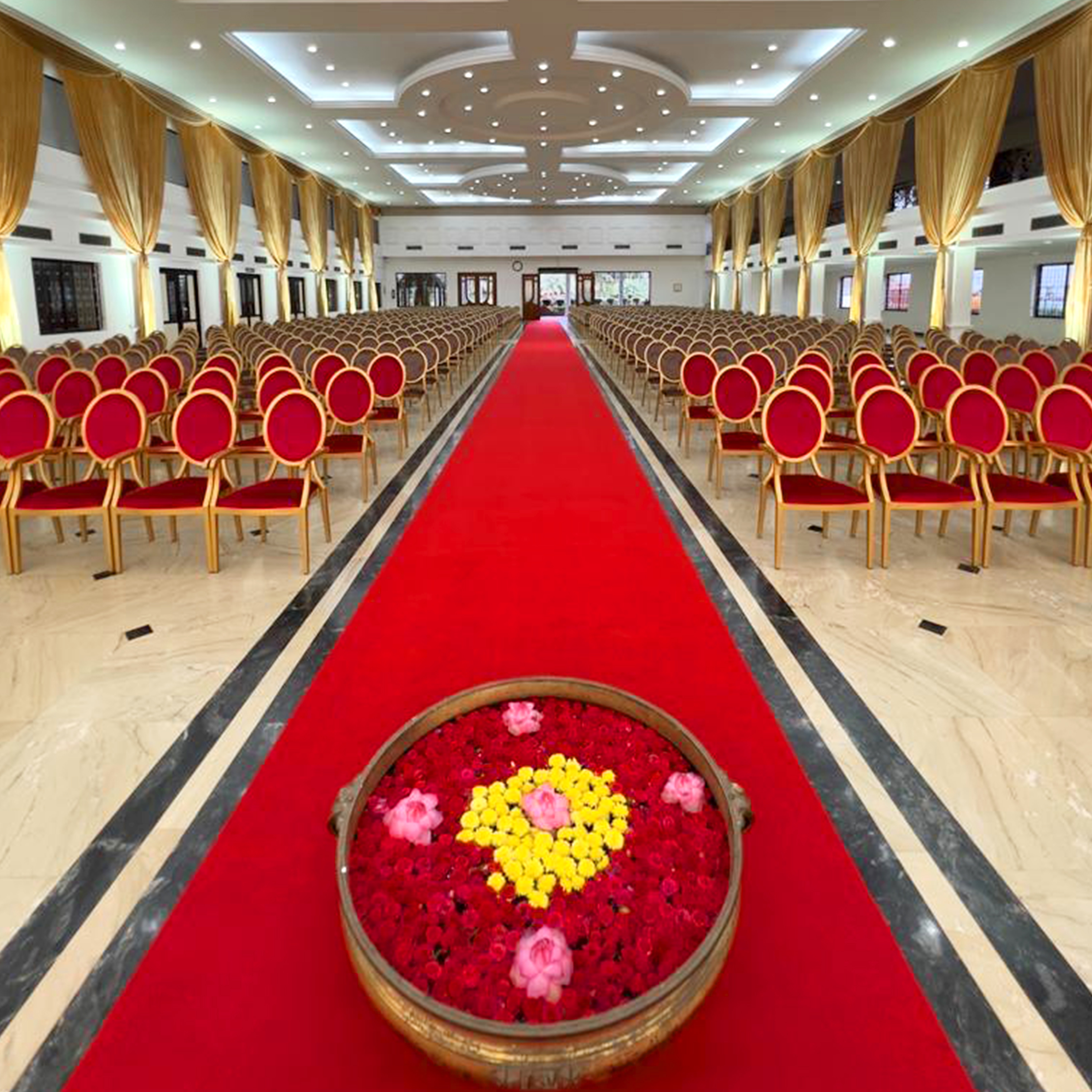 Large banquet hall with rows of red cushioned chairs, a red carpet aisle, and a circular floral arrangement at the front.