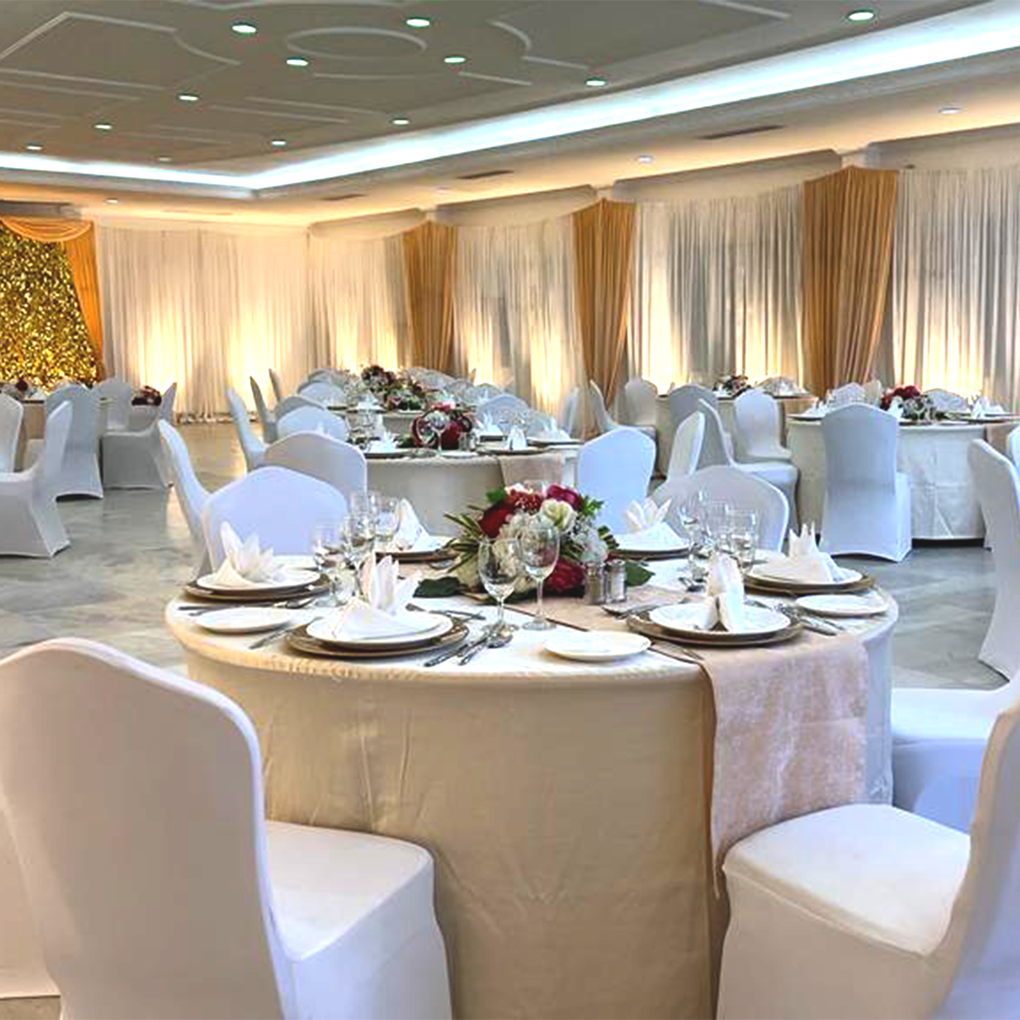 Elegant banquet hall with round tables dressed in ivory linens and white chairs, set with formal place settings and floral centerpieces.