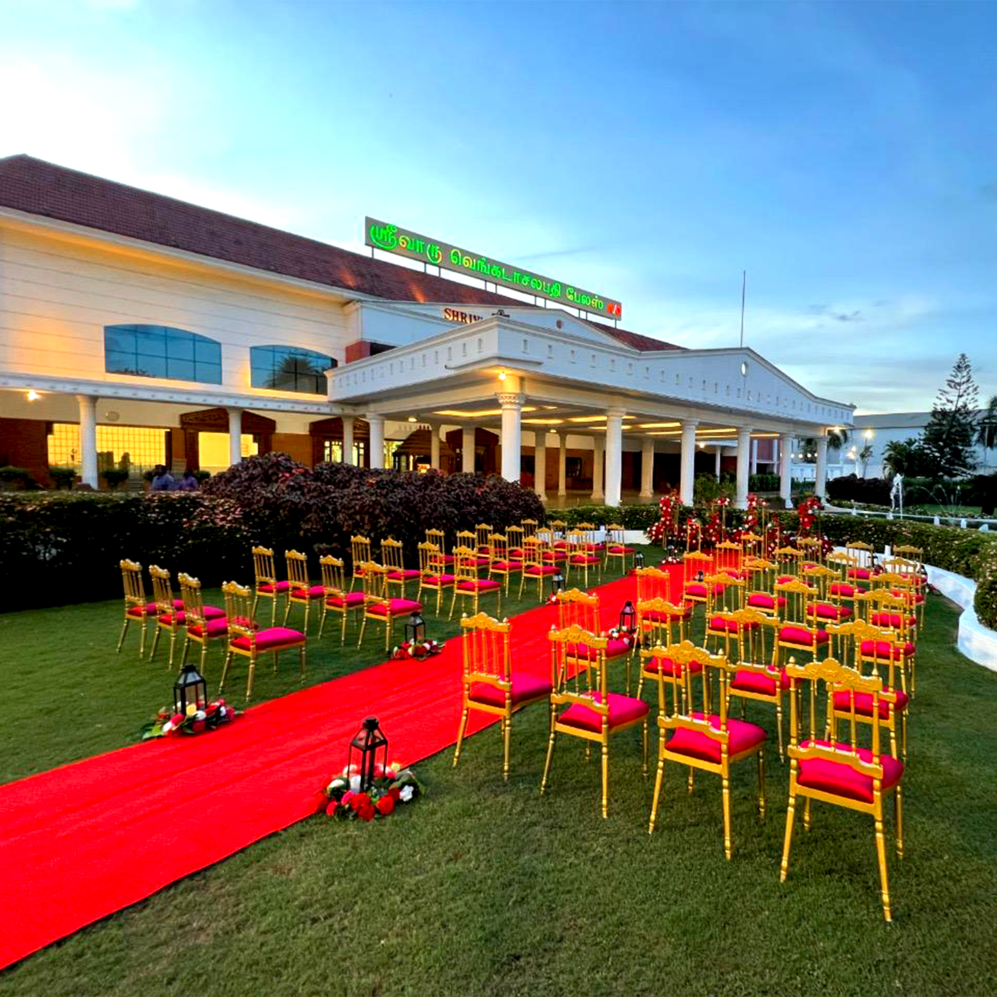 Outdoor wedding ceremony setup with golden chairs with red cushions arranged on green lawn, and a red carpet aisle decorated with lanterns and flowers.