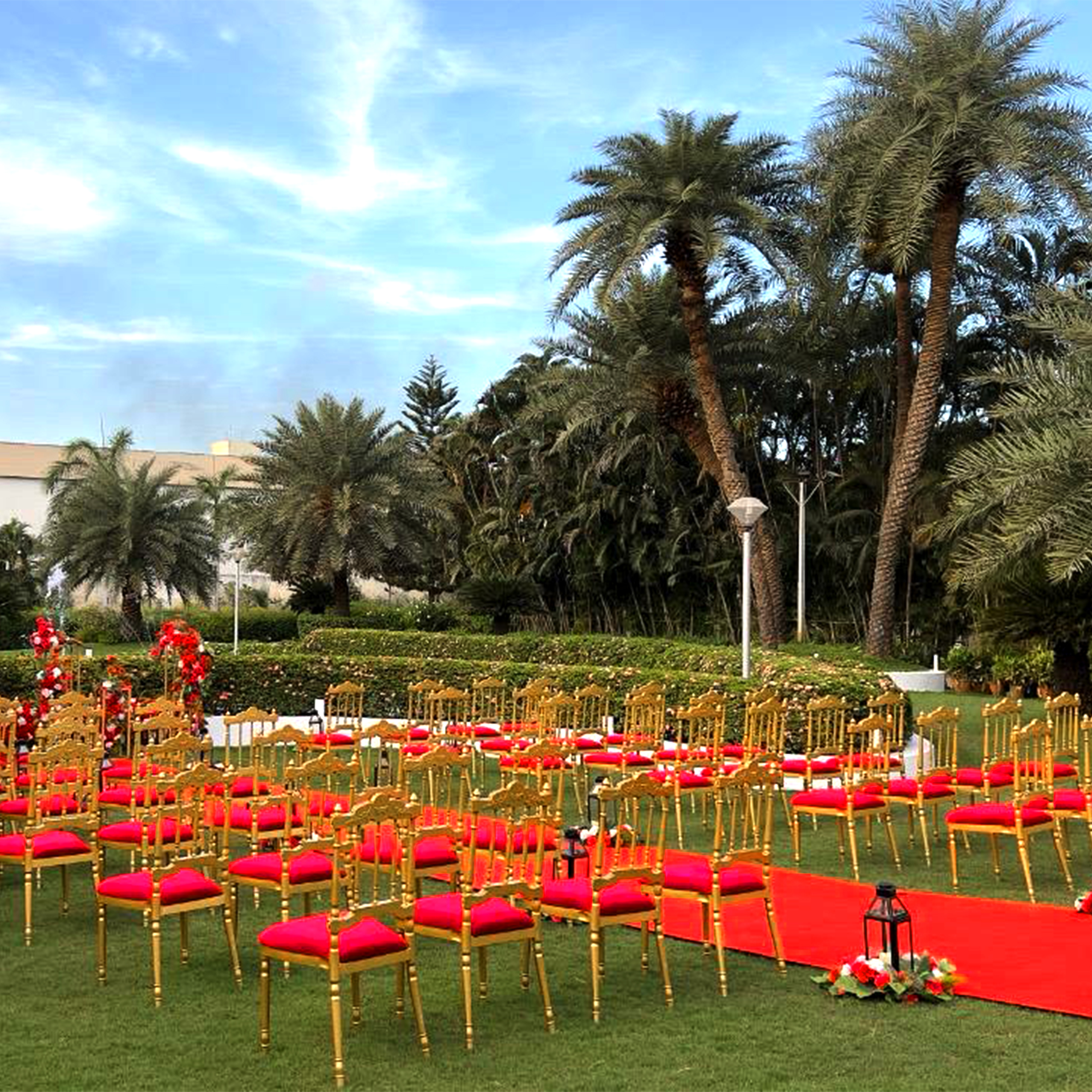 Outdoor event setup with gold chairs featuring red cushions arranged on grass with a red carpet aisle and tropical palm trees in the background.