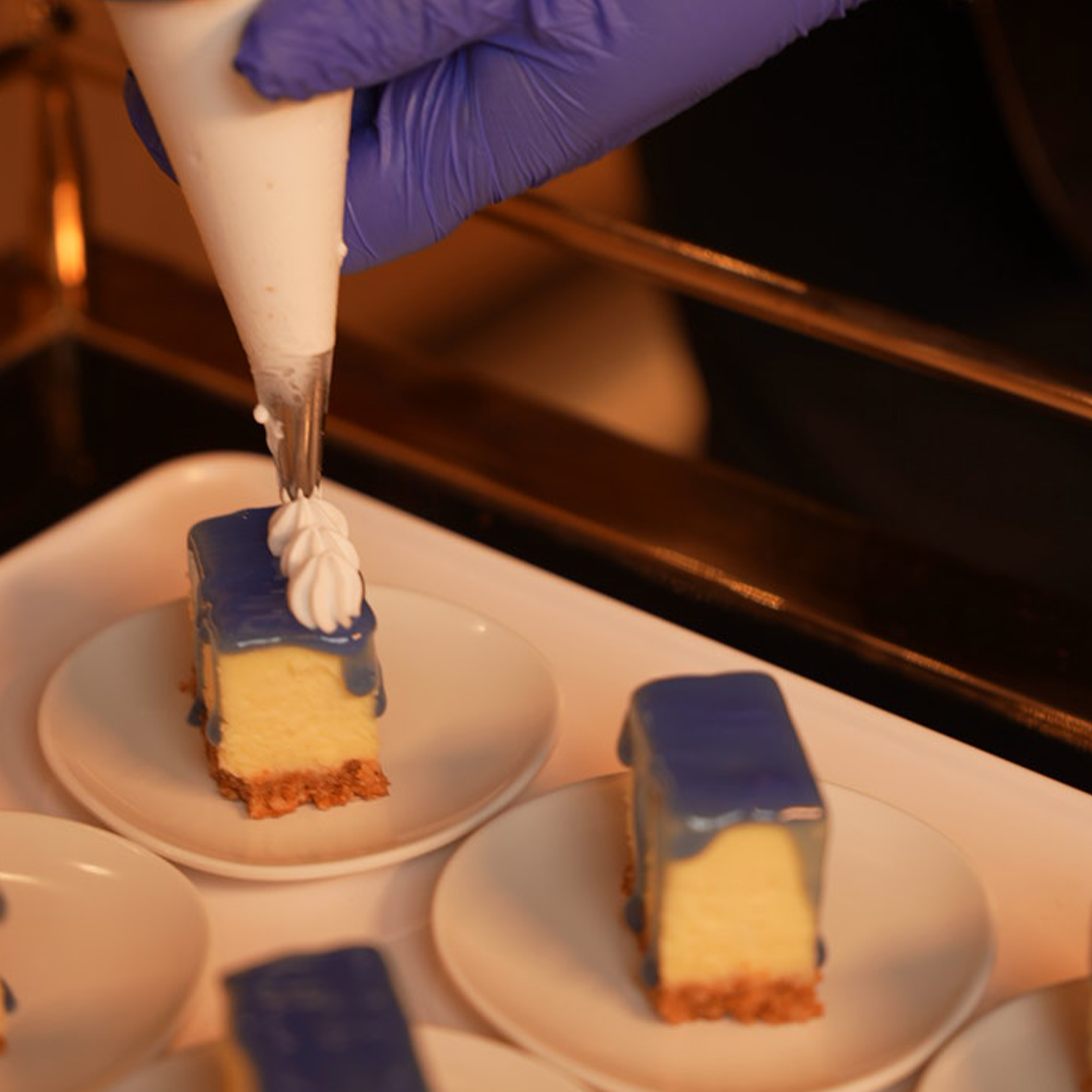 Hand with purple glove piping white cream decoration onto small rectangular blue-glazed cheesecake slices on white plates.