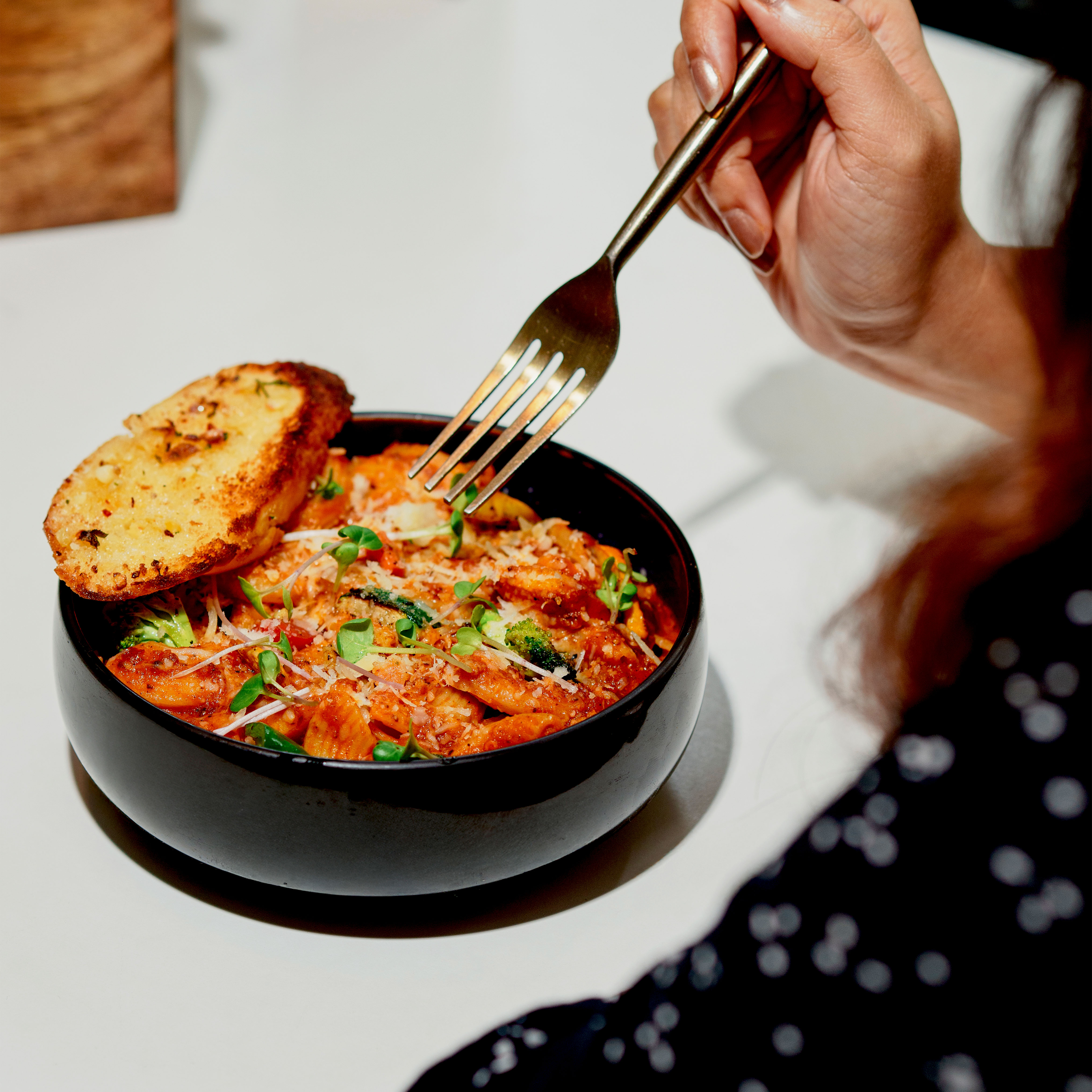 Person holding a fork poised to eat pasta with tomato sauce, garnished with greens and cheese, served with a slice of garlic bread in a black bowl.