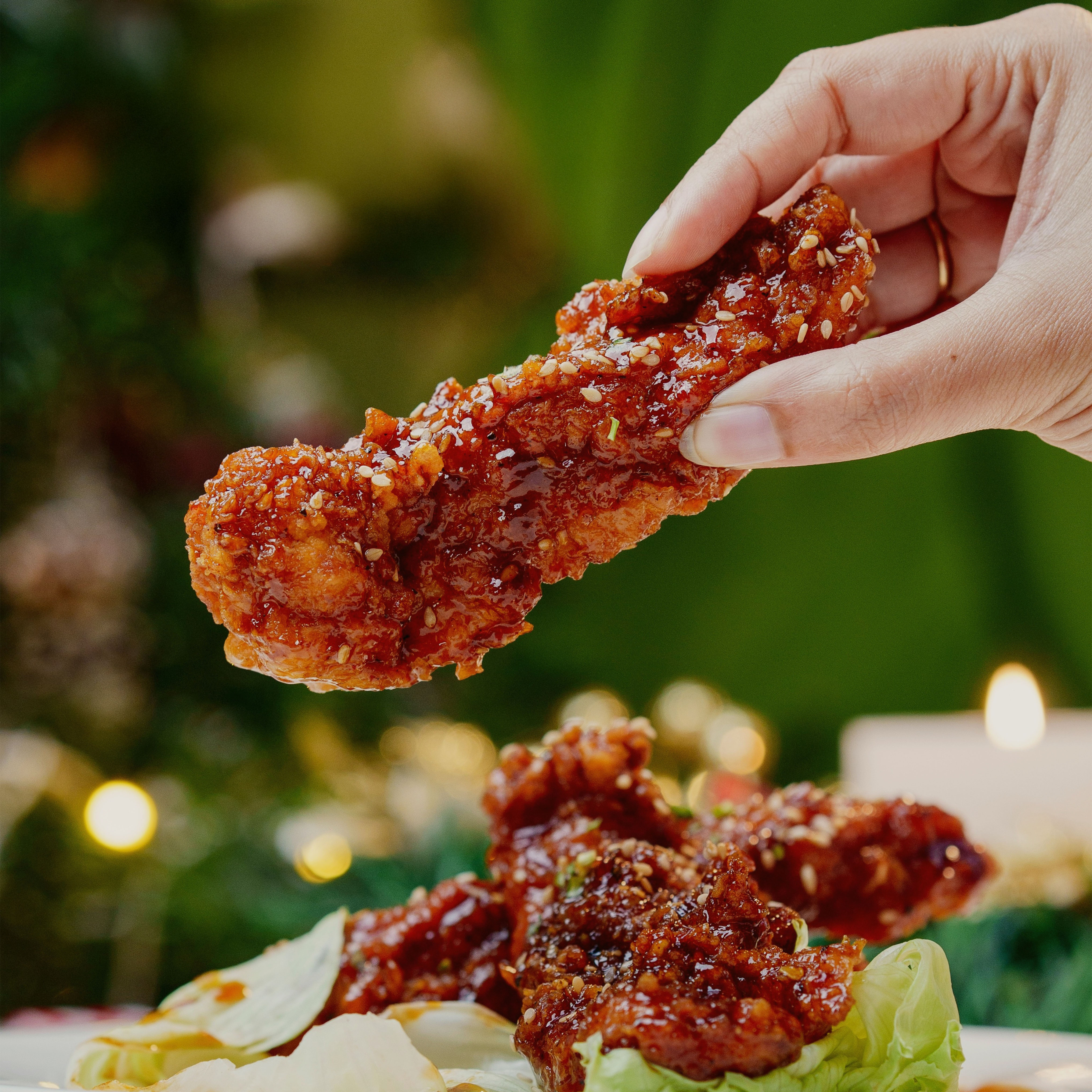 Hand holding a piece of glazed crispy fried chicken garnished with sesame seeds over a plate with lettuce and more fried chicken pieces.