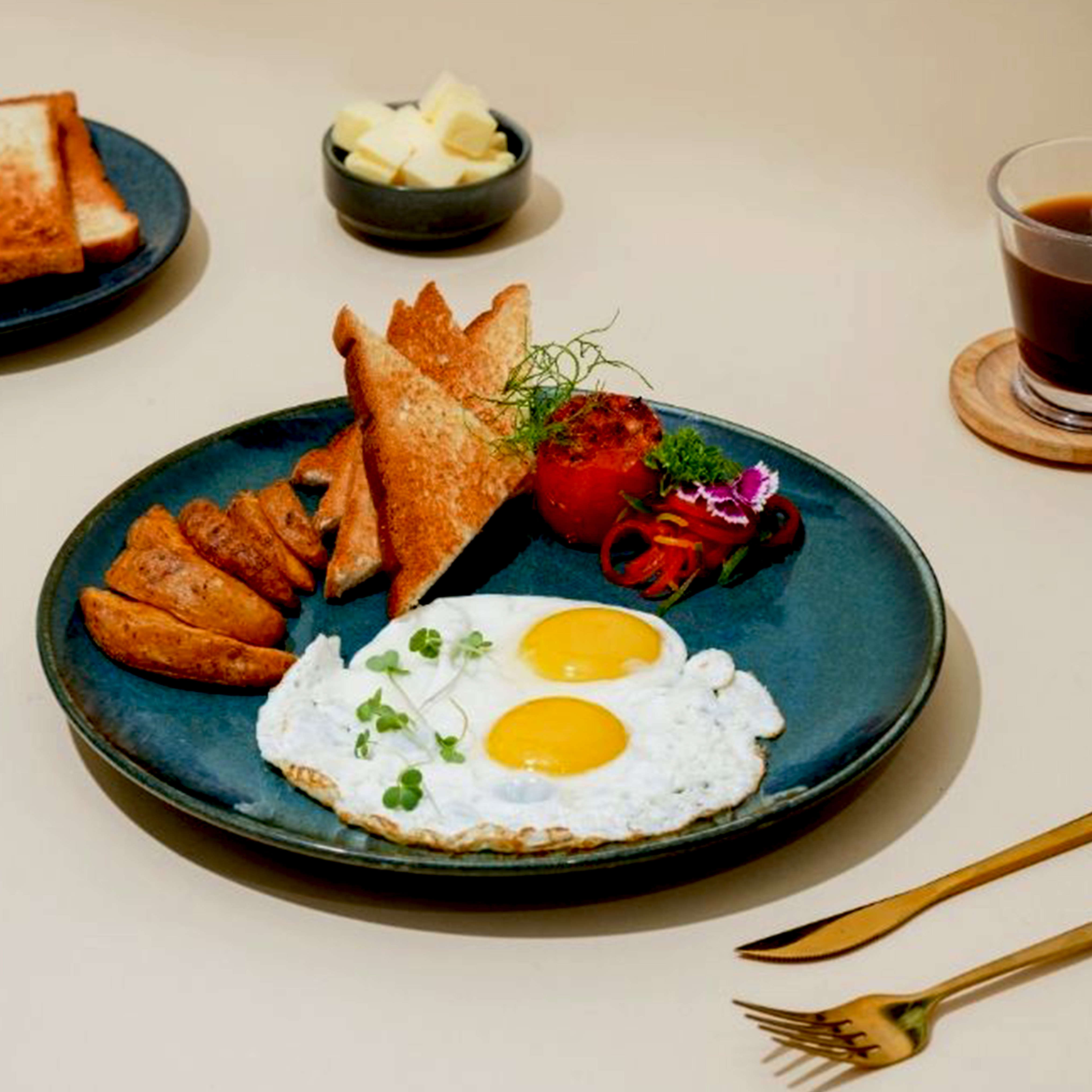 Plate with sunny side up eggs, toast slices, potato wedges, grilled tomato, and garnish accompanied by a glass of coffee and a small bowl of butter cubes.