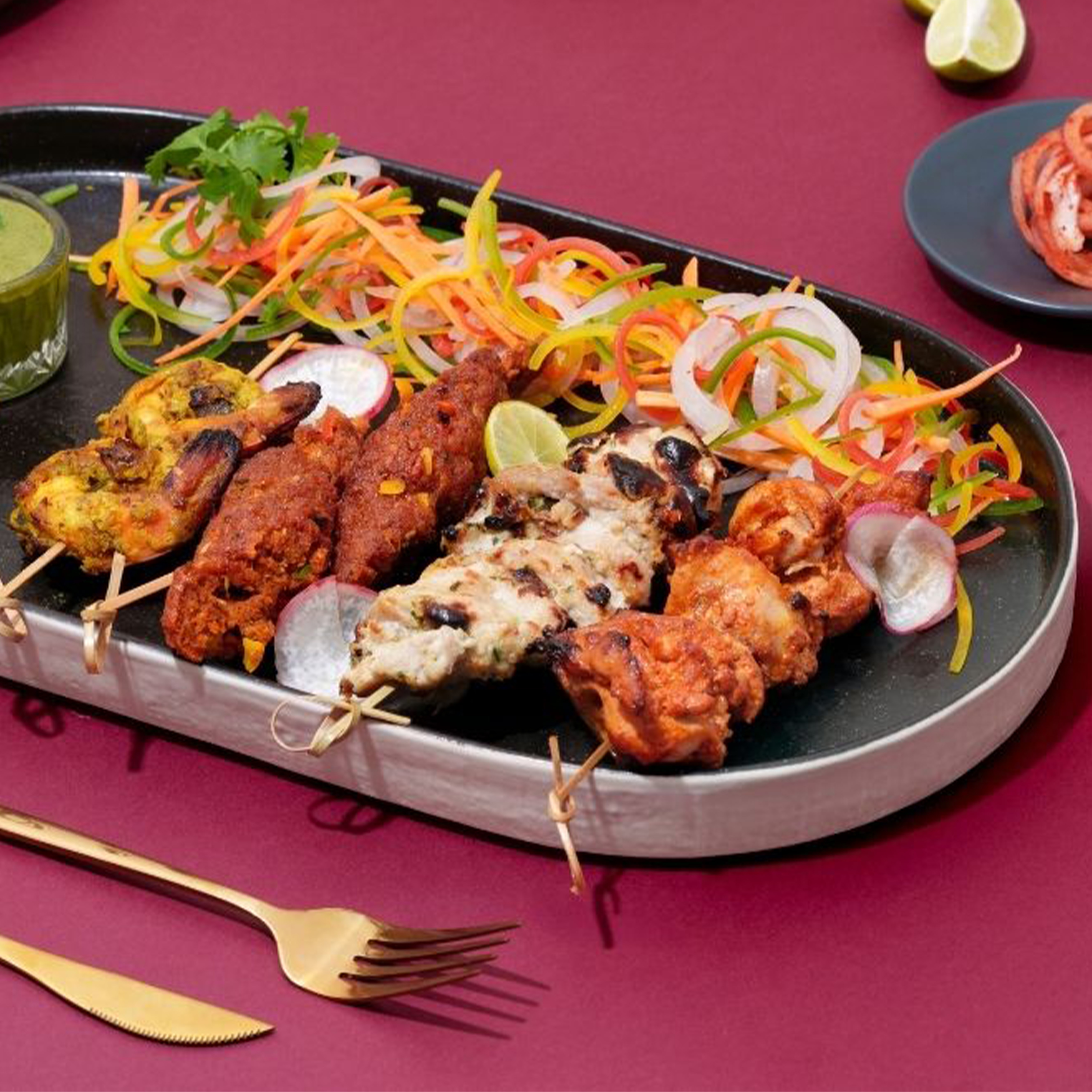 Assorted grilled non-vegetarian tandoori skewers with sliced onions, carrots, and lemon on a black platter with green chutney on the side.