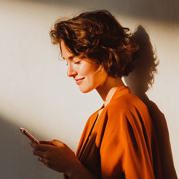 Smiling woman in an orange top looking at her smartphone with sunlight casting her shadow on the wall.