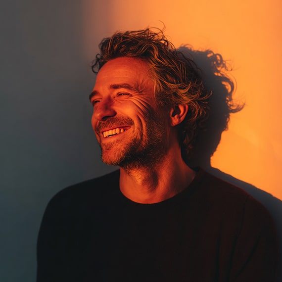 Smiling man with curly hair and beard lit by warm orange light casting a shadow on the wall.