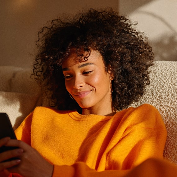 Smiling woman with curly hair wearing an orange sweater, sitting on a beige couch and looking at her phone.