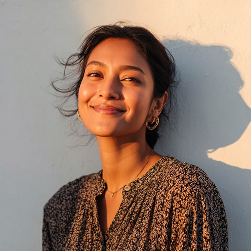 Smiling woman with dark hair wearing gold hoop earrings and a patterned blouse standing against a light wall with a shadow.