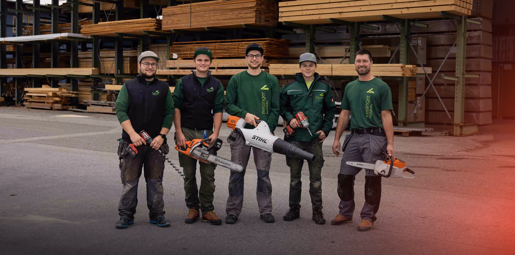 Fünf Männer in Arbeitskleidung stehen vor einem Holzlager und halten Werkzeuge wie Motorsägen, Bohrer und Laubbläser.