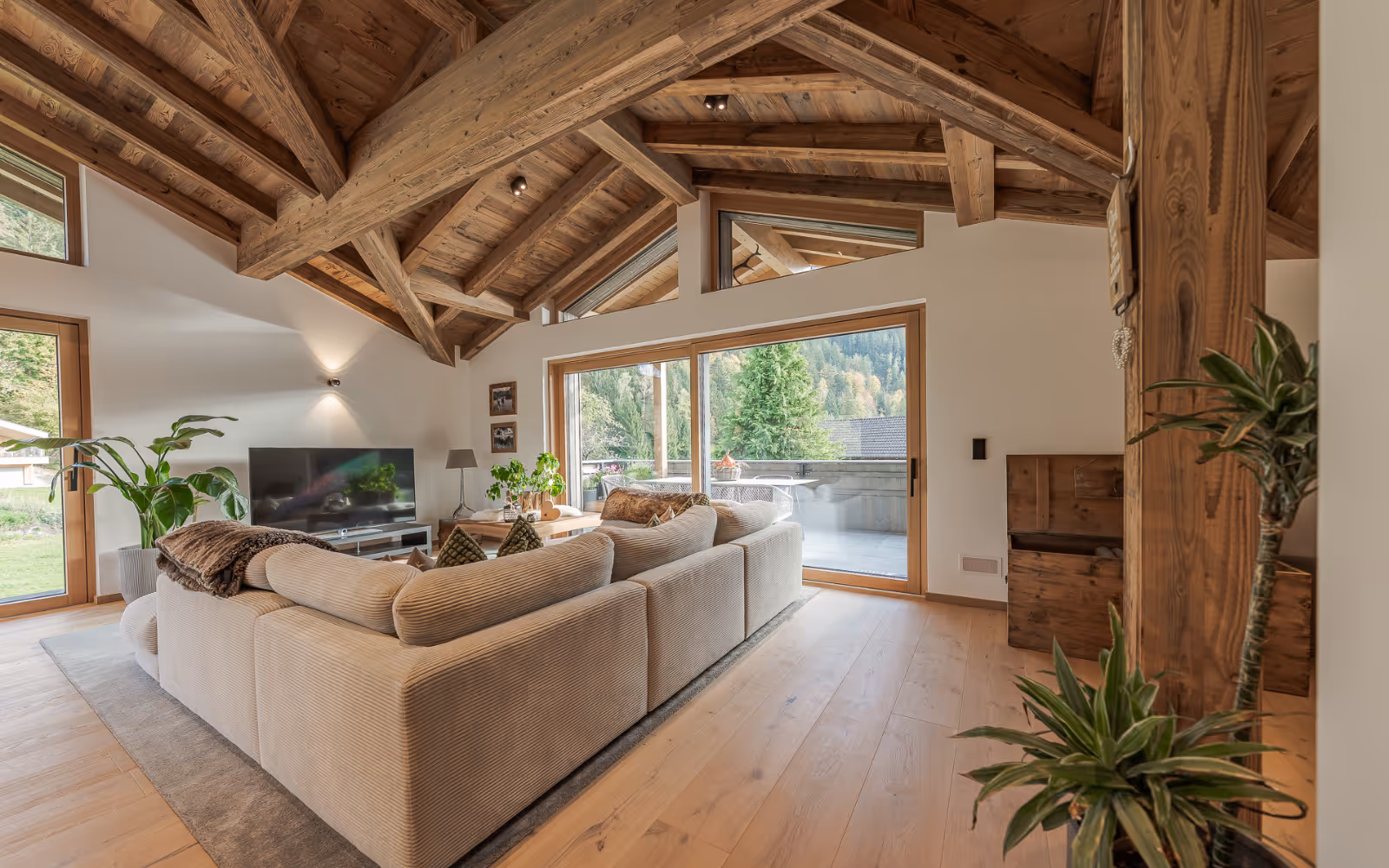 Modernes Wohnzimmer mit großer Creme-eckcouch, Holzbalkendecke, Fernseher, Pflanzen und großen Fenstern mit Blick auf eine Terrasse und grüne Berge.