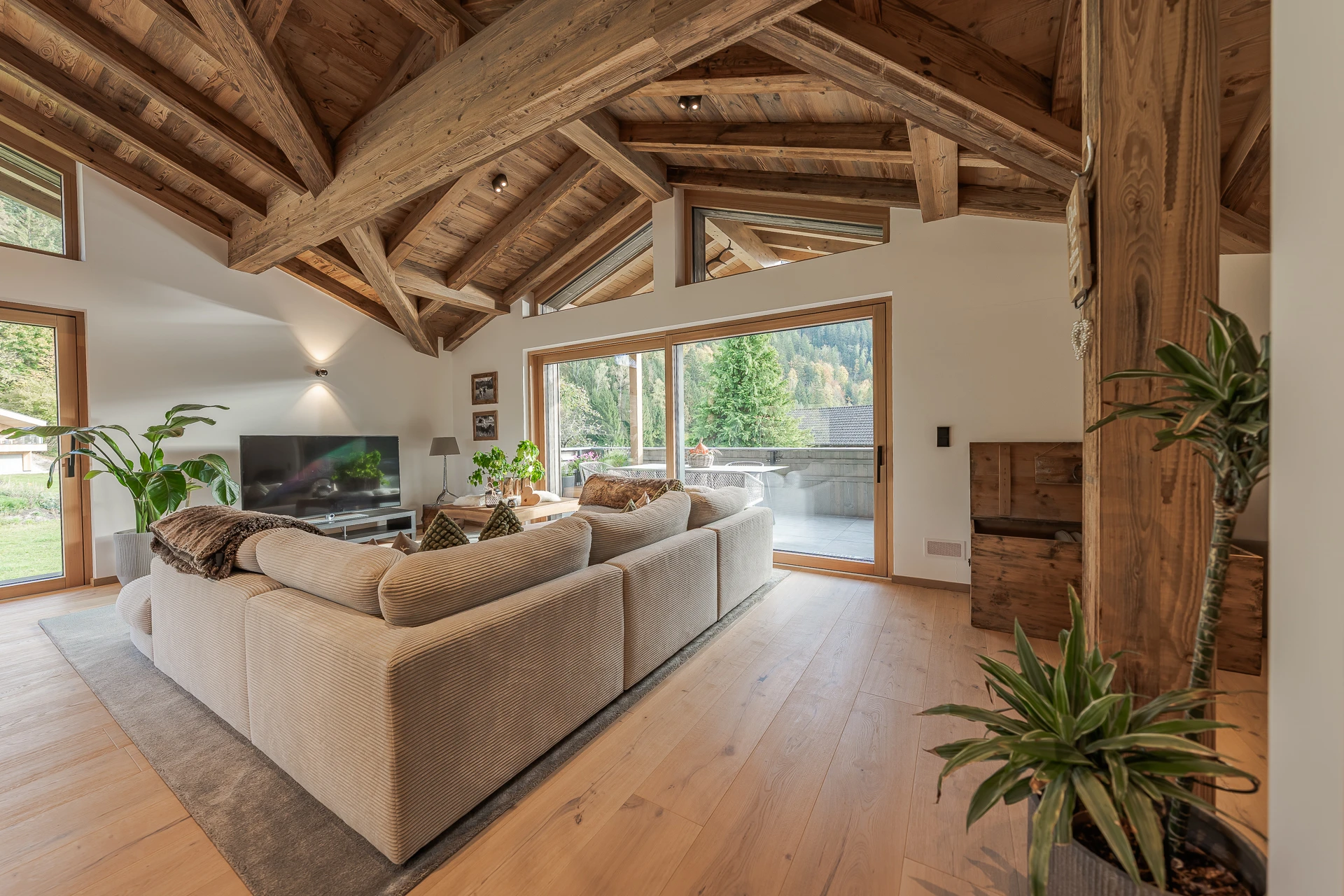 Modernes Wohnzimmer mit großer Creme-eckcouch, Holzbalkendecke, Fernseher, Pflanzen und großen Fenstern mit Blick auf eine Terrasse und grüne Berge.