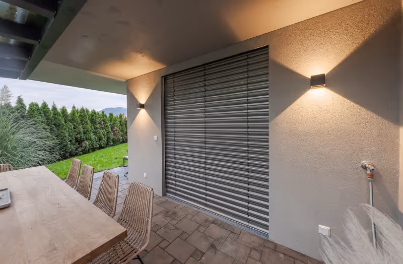 Überdachte Terrasse mit großem Esstisch aus Holz und geflochtenen Stühlen, angrenzend an eine geschlossene Außenjalousie an der Hauswand. Warmes Licht von zwei Wandlampen erzeugt eine gemütliche Atmosphäre, während der Blick ins Grüne Ruhe und Natur vermittelt.