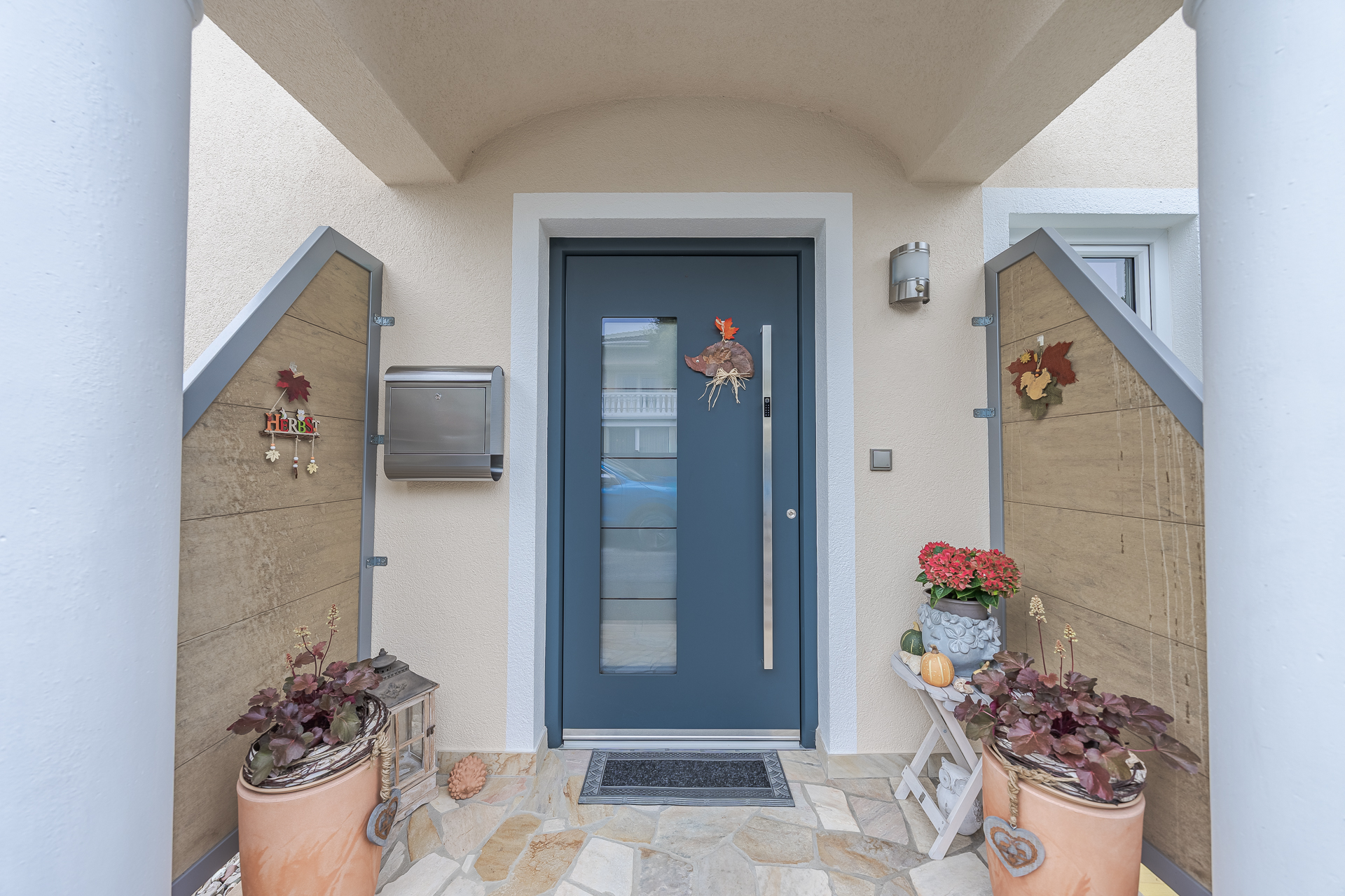 Moderne Haustür in dunkelblau mit schmalem Glas­einsatz und vertikalem Edelstahlgriff, eingerahmt von einer hellen Fassade. Der Eingangsbereich ist herbstlich dekoriert mit Blumen, Kürbissen und Pflanzenarrangements und wirkt einladend und gepflegt.