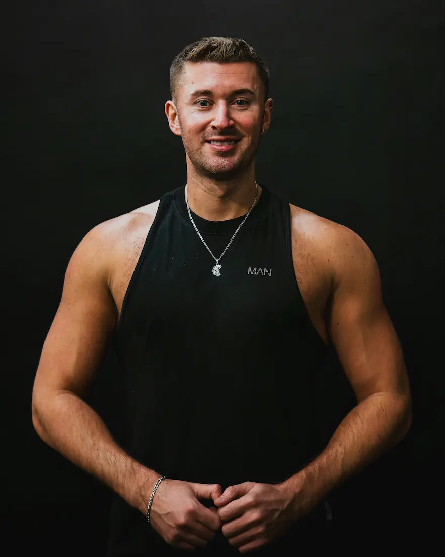 Smiling muscular man wearing a black sleeveless shirt and silver necklace against a black background.