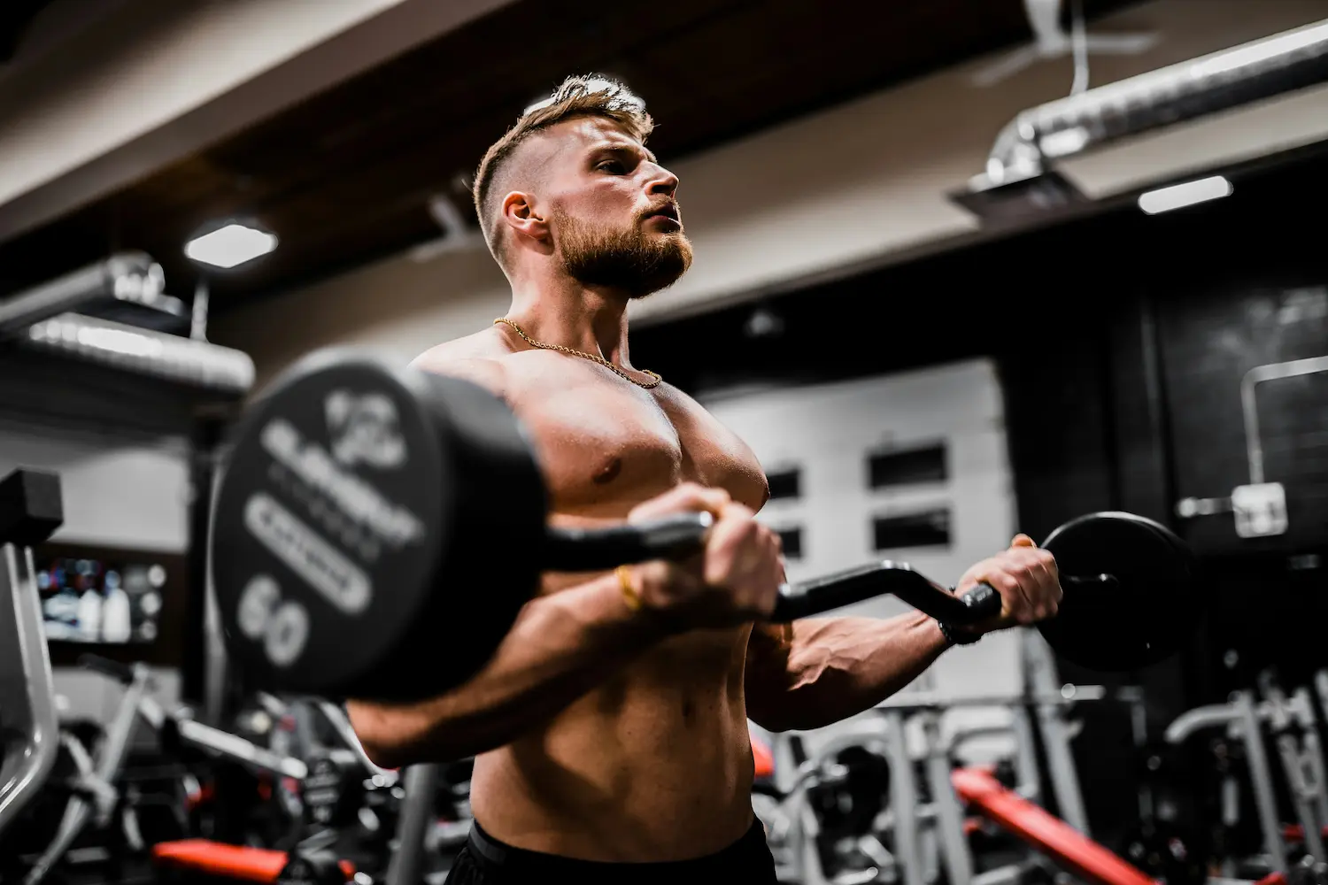 Muscular shirtless man performing barbell curls in a gym.