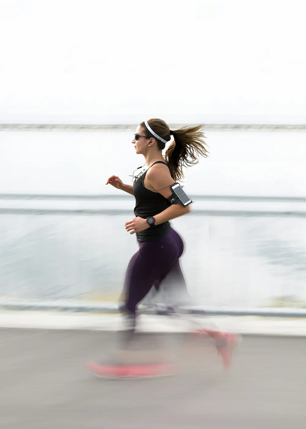 Woman jogging by the water wearing sunglasses, earbuds, and a smartphone armband.