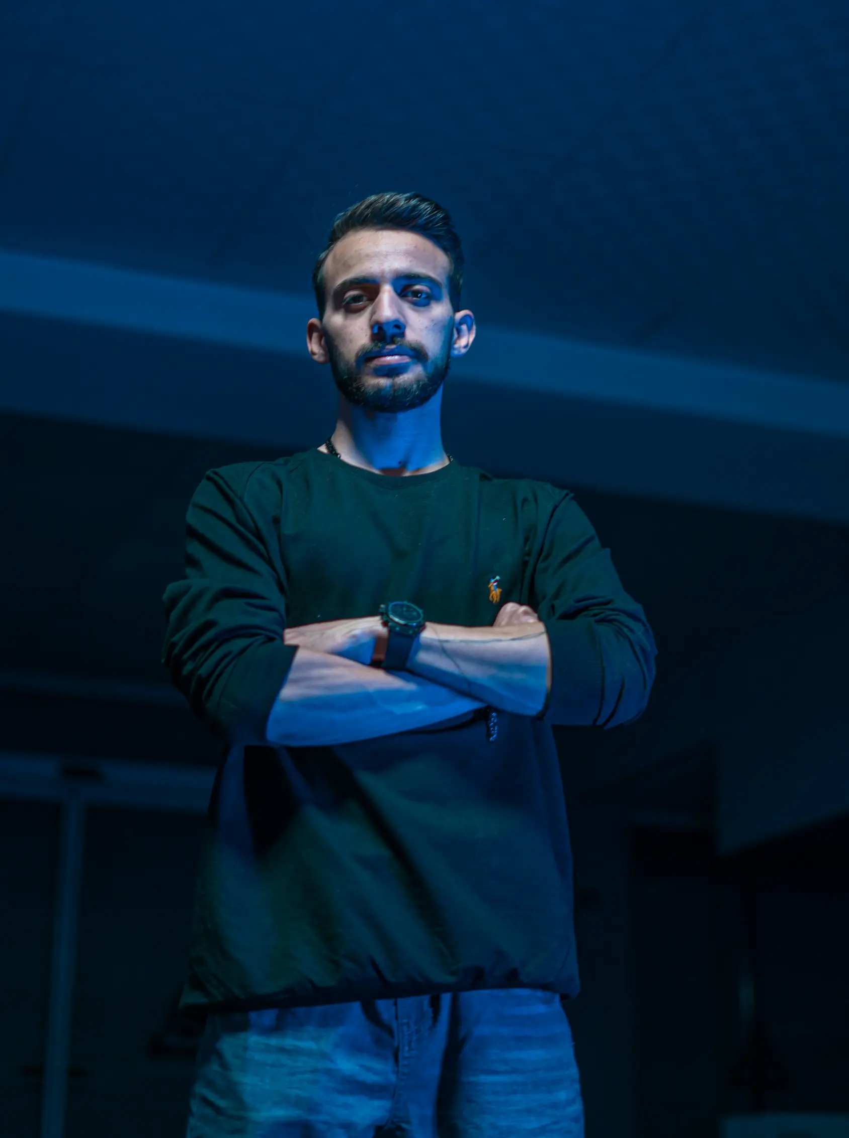 Young man with crossed arms wearing a dark long-sleeve shirt and jeans, illuminated by blue lighting.