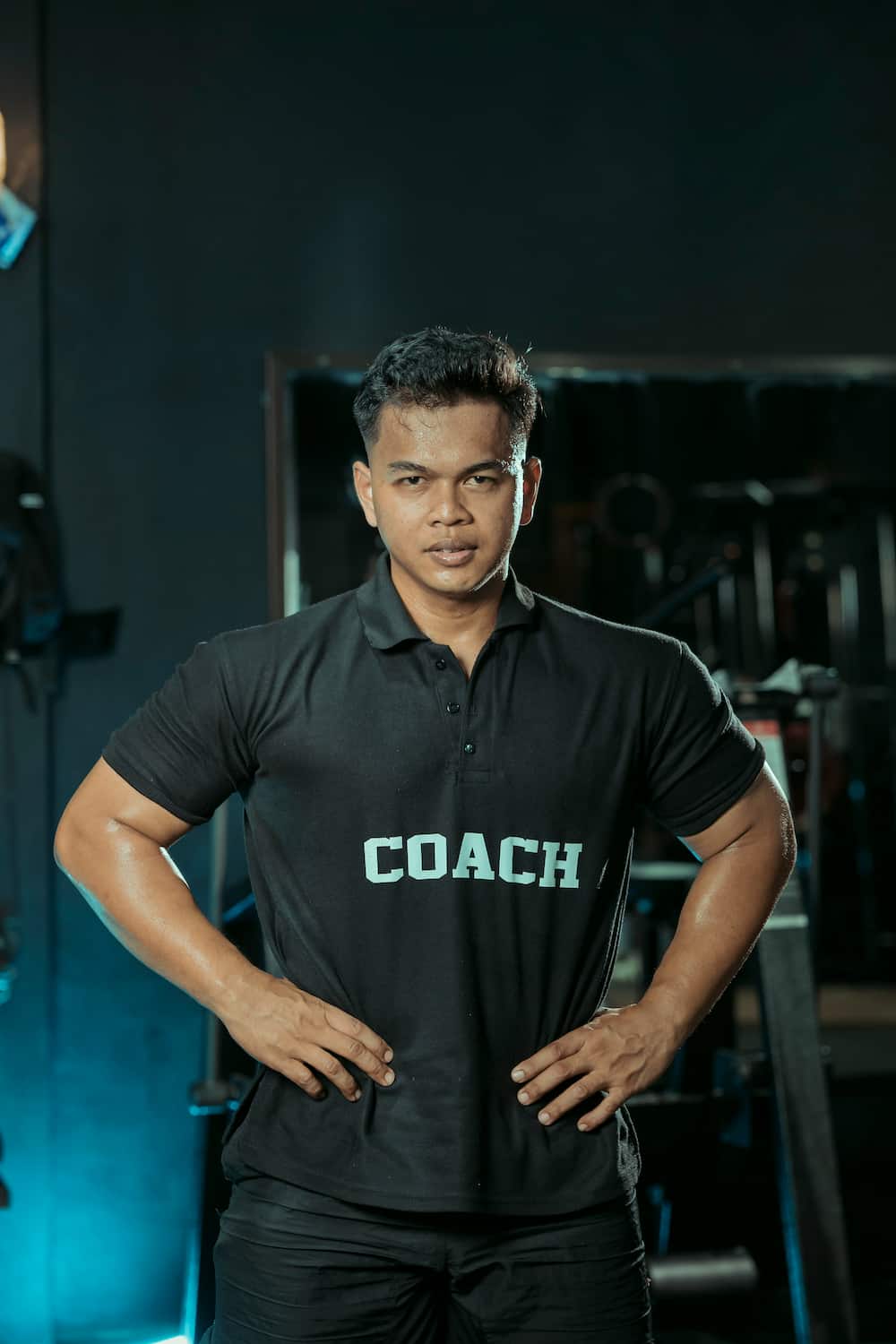 Fit man wearing a black polo shirt with the word 'COACH' printed on it, standing with hands on hips in a gym.