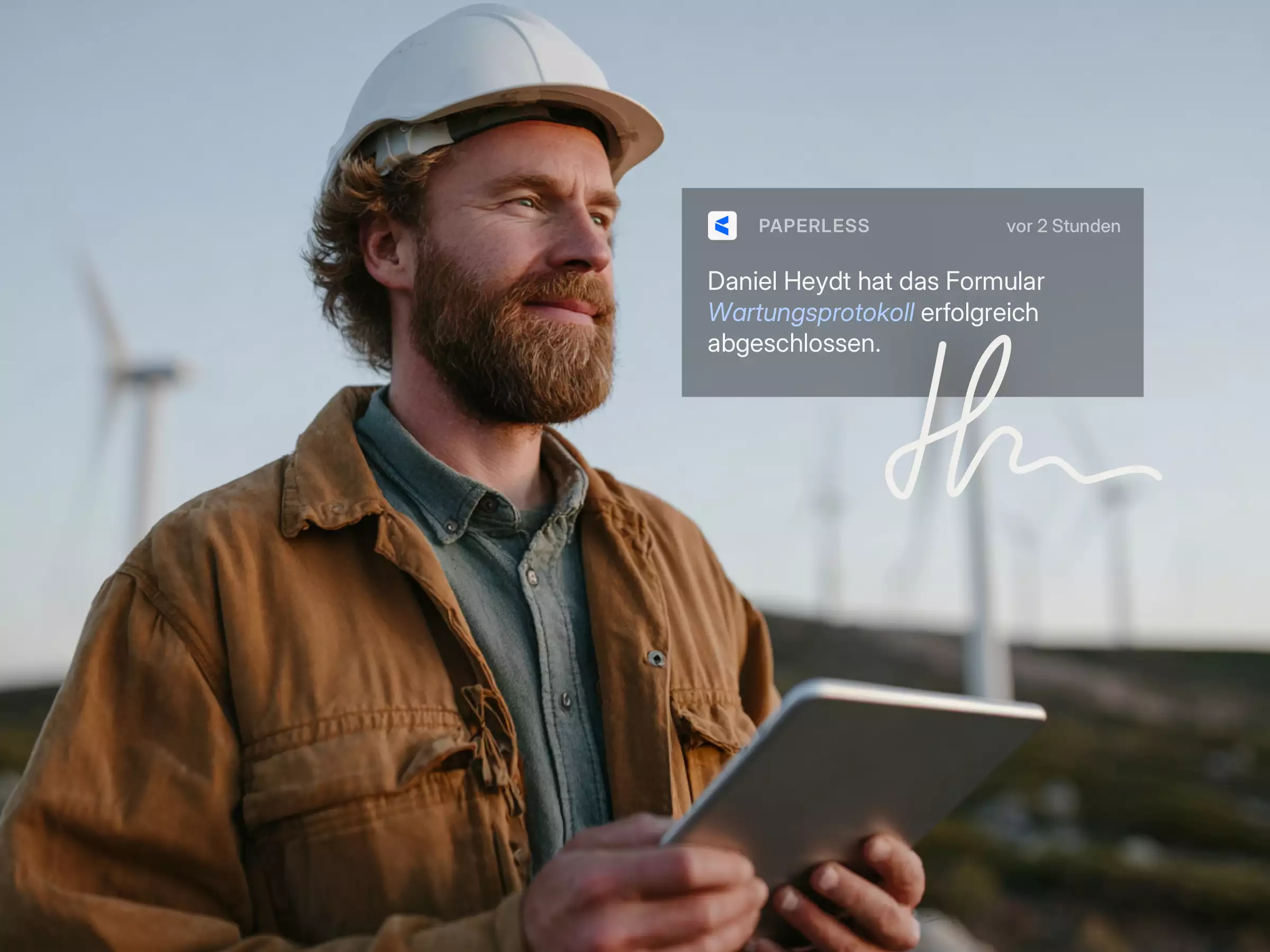 Ein Mann mit Schutzhelm und brauner Jacke hält ein Tablet vor Windkraftanlagen bei Sonnenuntergang.