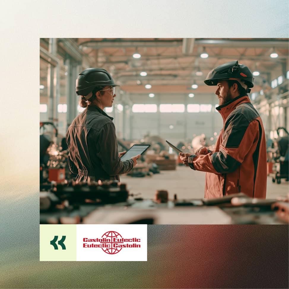Zwei Industriearbeiter in Schutzhelmen unterhalten sich in einer großen, gut beleuchteten Fabrikhalle, während sie Tablets in der Hand halten.