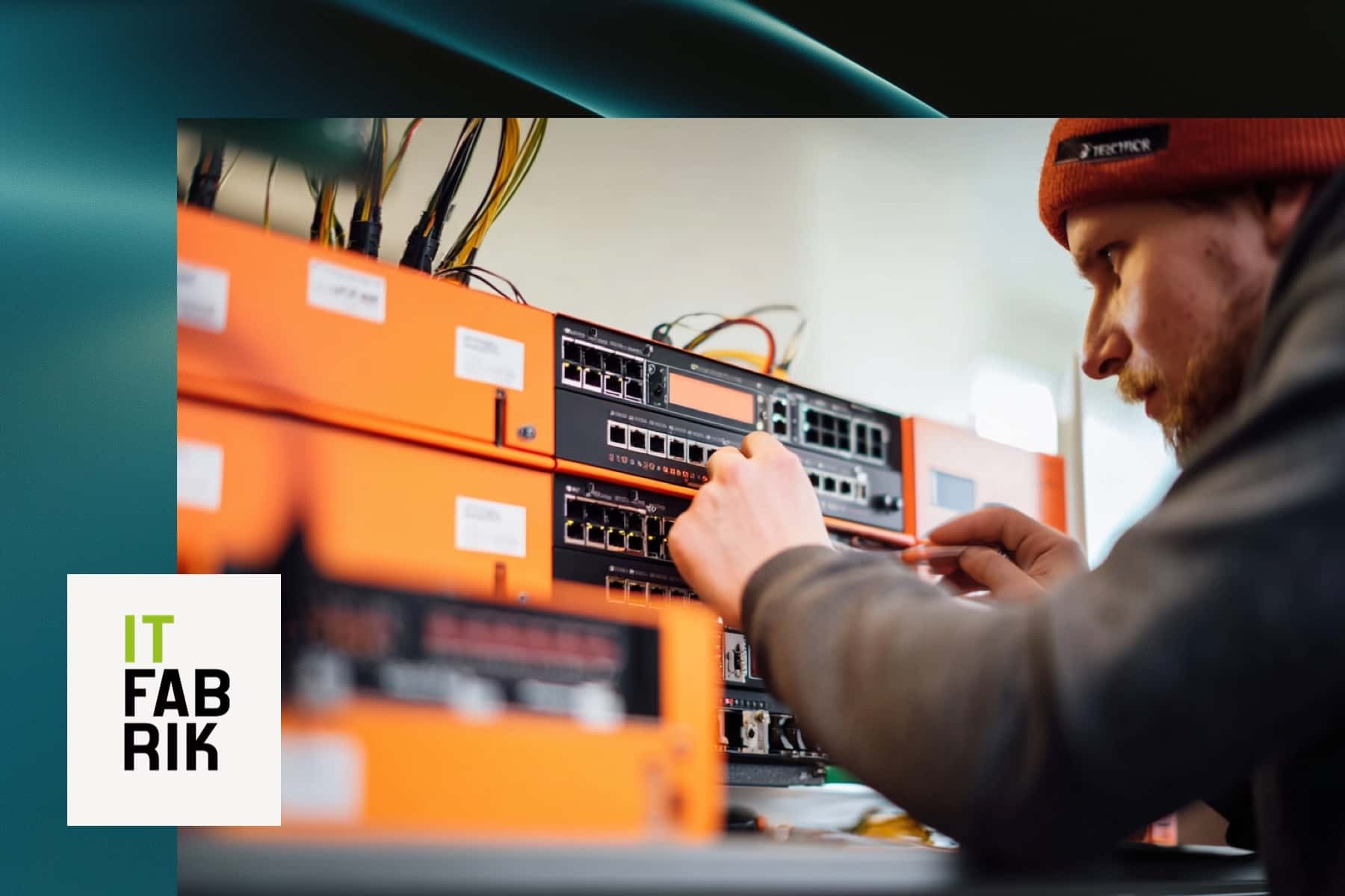 Ein Techniker mit Mütze und Bart steckt ein Netzwerkkabel in einen orangefarbenen Serverschrank mit mehreren Switches und Patchfeldern. Unten links das Logo der IT Fabrik.