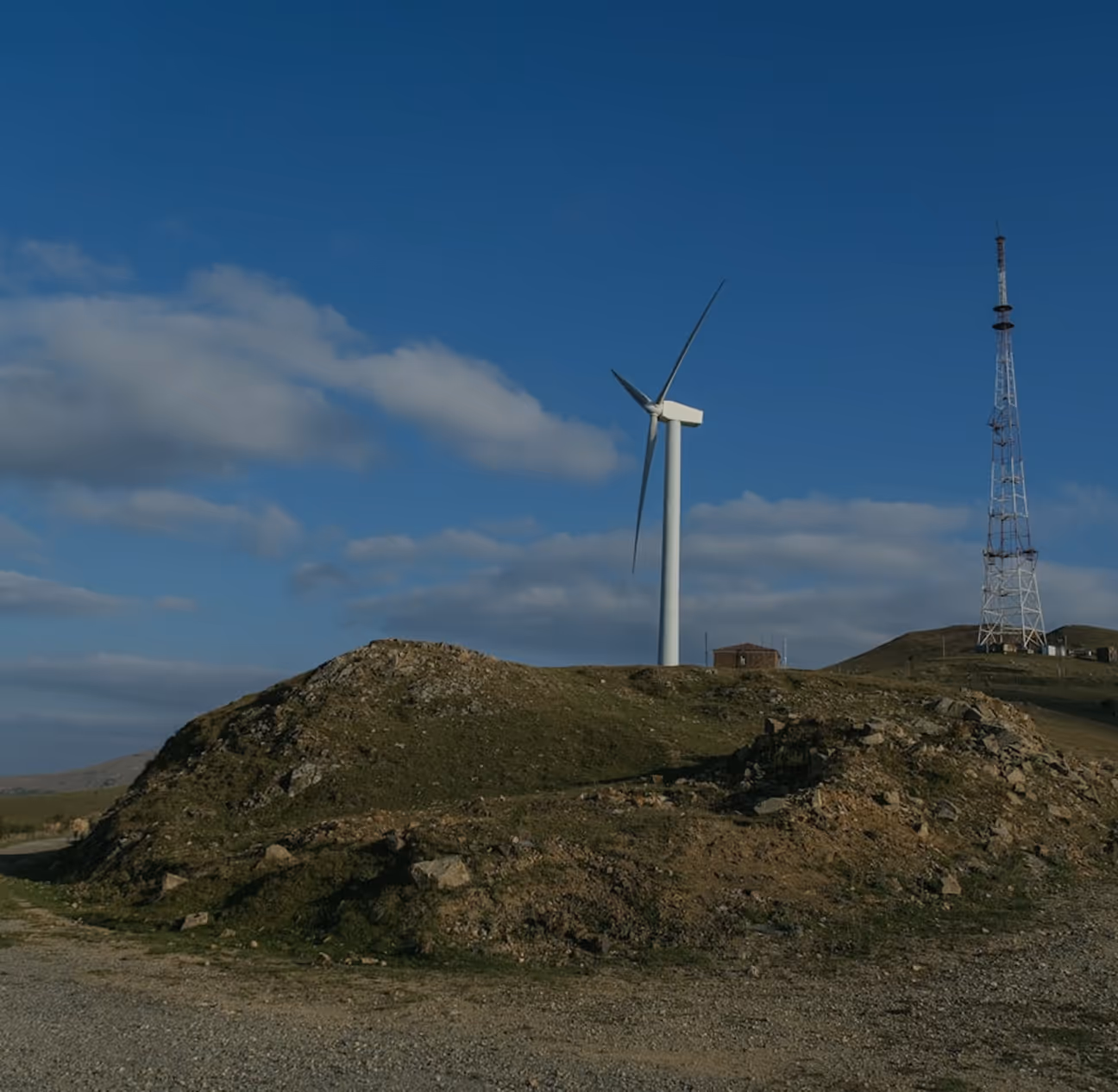 Wind turbine and telecommunications tower on a rocky hill under a blue sky, representing renewable energy generation and secure grid communications for utility companies and grid operators.