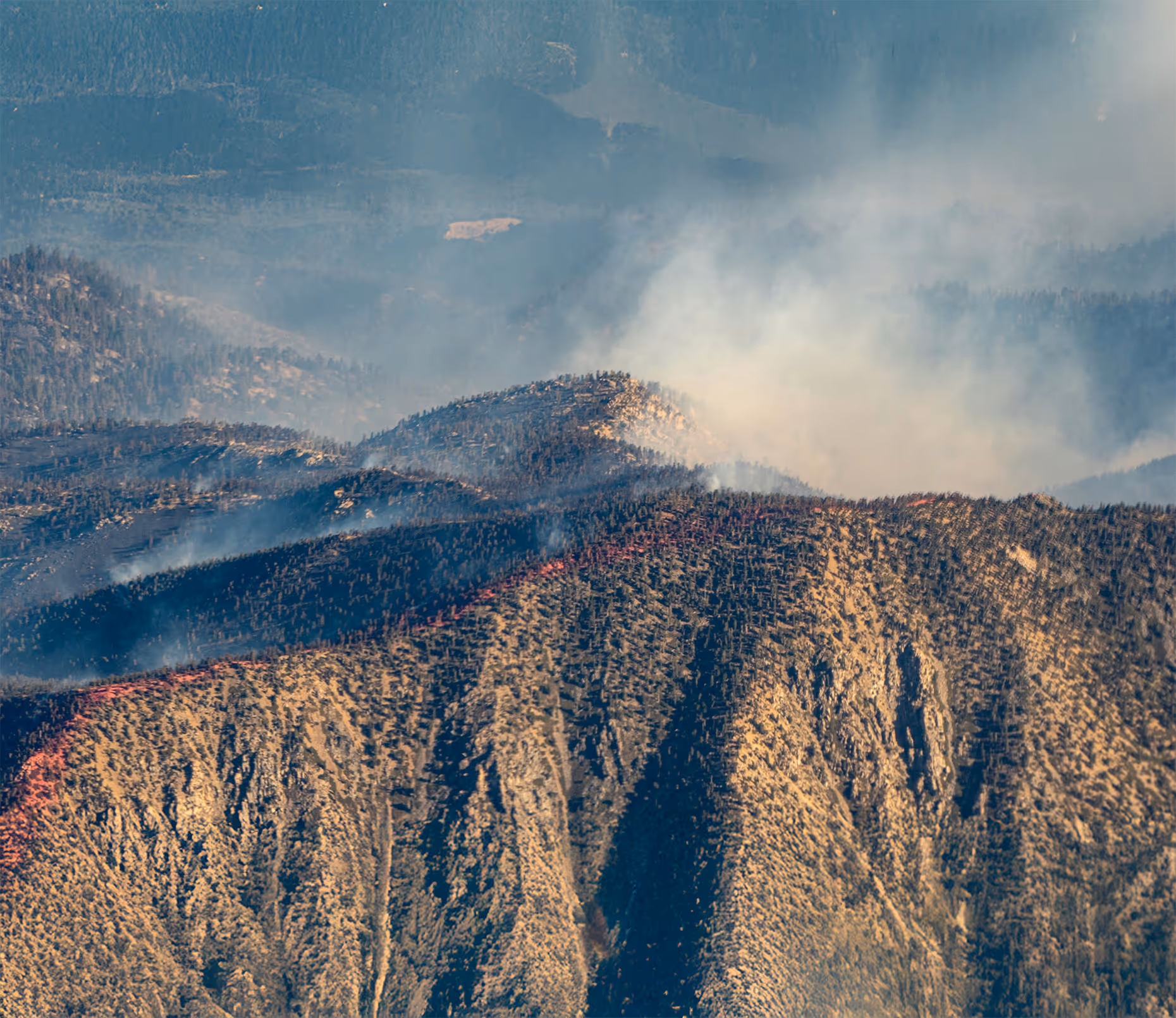 Aerial view of a forested mountain range with smoke rising from a wildfire, representing emergency management and national security operations for disaster response, environmental monitoring, and threat detection.