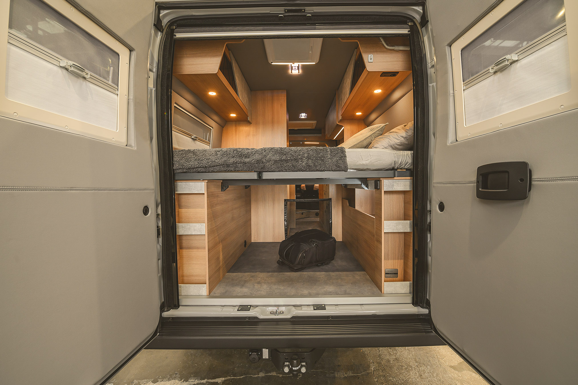 View into a camper van from the rear with an elevated bed, wooden cabinetry, and a black backpack on the floor underneath.