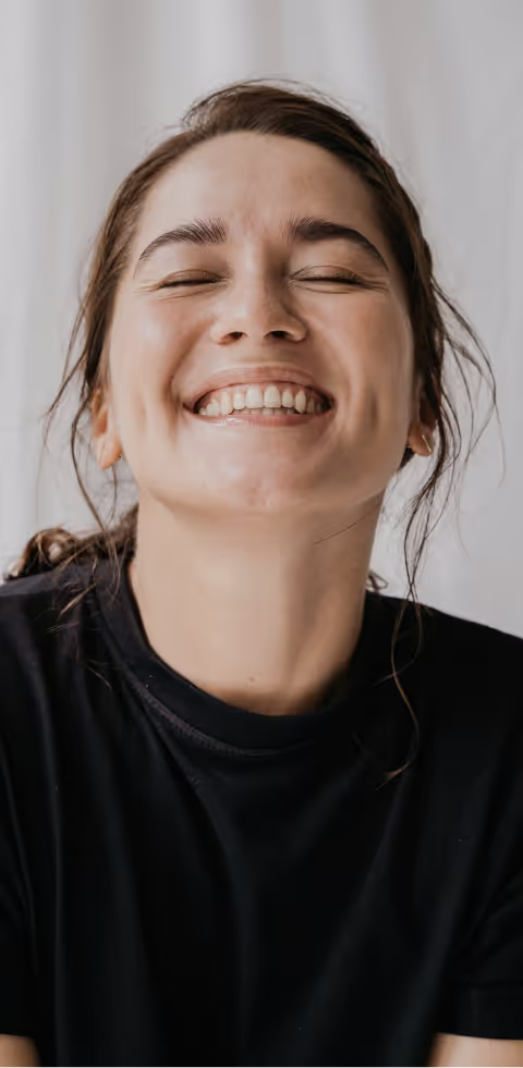 Close-up of a joyful woman in a black t-shirt, smiling brightly with her eyes closed, standing against a soft white backdrop