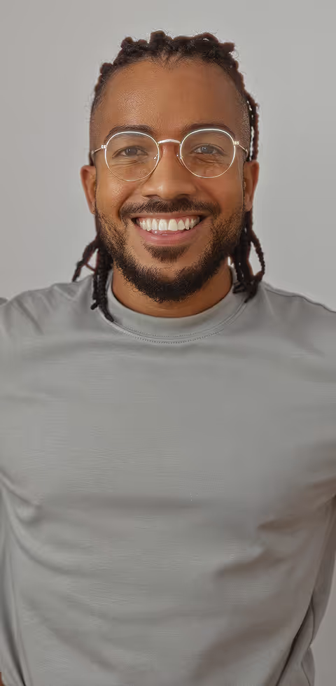 Smiling man with braided hair and a beard, wearing round glasses and a light gray t-shirt, standing in front of a plain light gray background