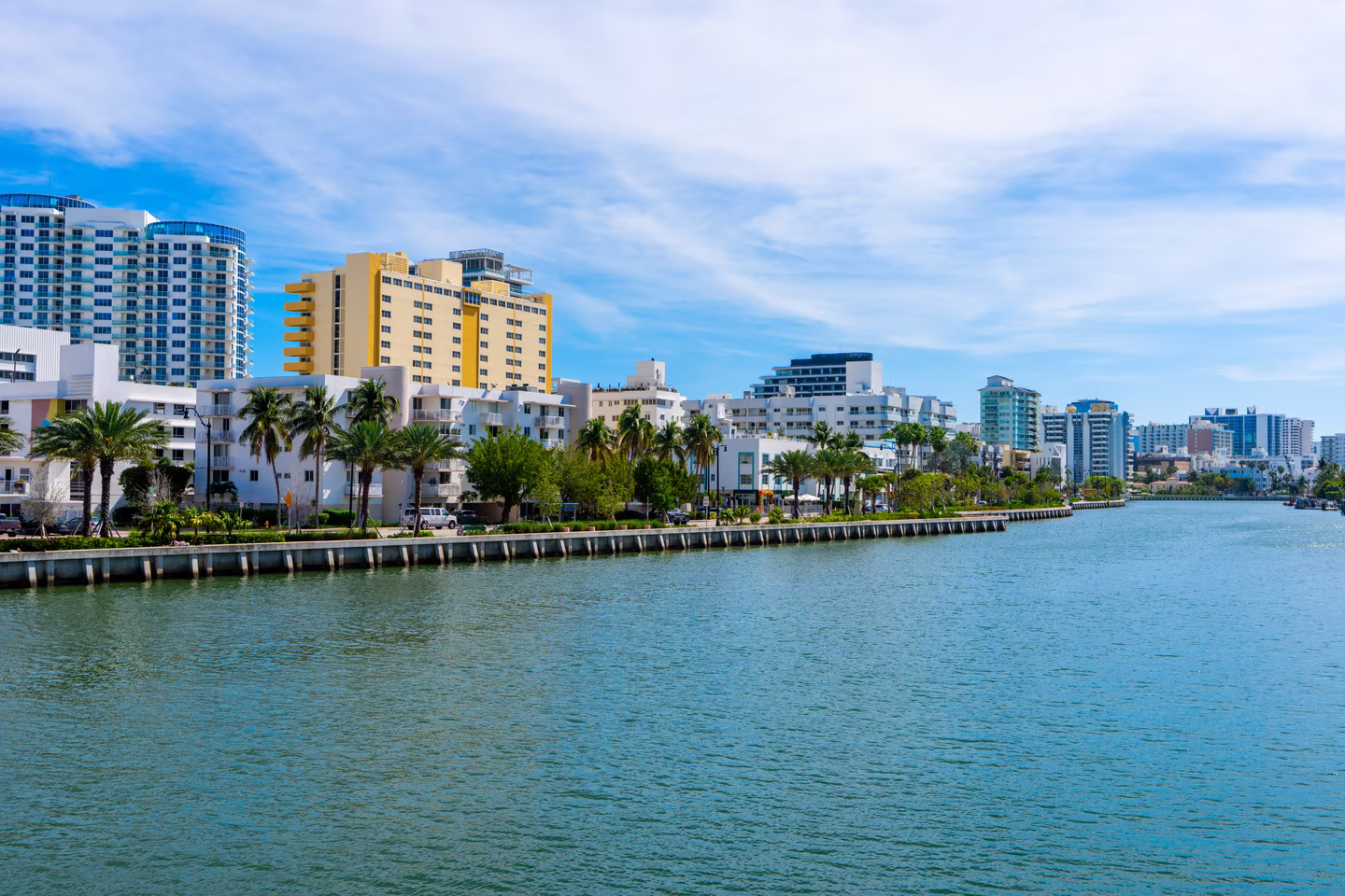 A scenic waterfront view of Miami showcases the calm waters of a river in the foreground, reflecting the bright blue sky. Along the riverbank, a row of modern buildings rises, including white and yellow high-rise condos with glass windows, typical of Miami's architectural style. Lush palm trees and manicured greenery line the waterfront, adding a tropical vibe. In the background, taller skyscrapers dot the skyline under a partly cloudy sky, capturing the vibrant, urban energy of this iconic coastal city.
