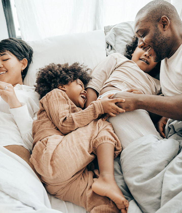 A family of four is lying on a bed, smiling and laughing together. They are dressed in cozy, neutral-toned clothing, and the scene is set against a backdrop of soft white bedding and curtains, creating a warm and intimate atmosphere.