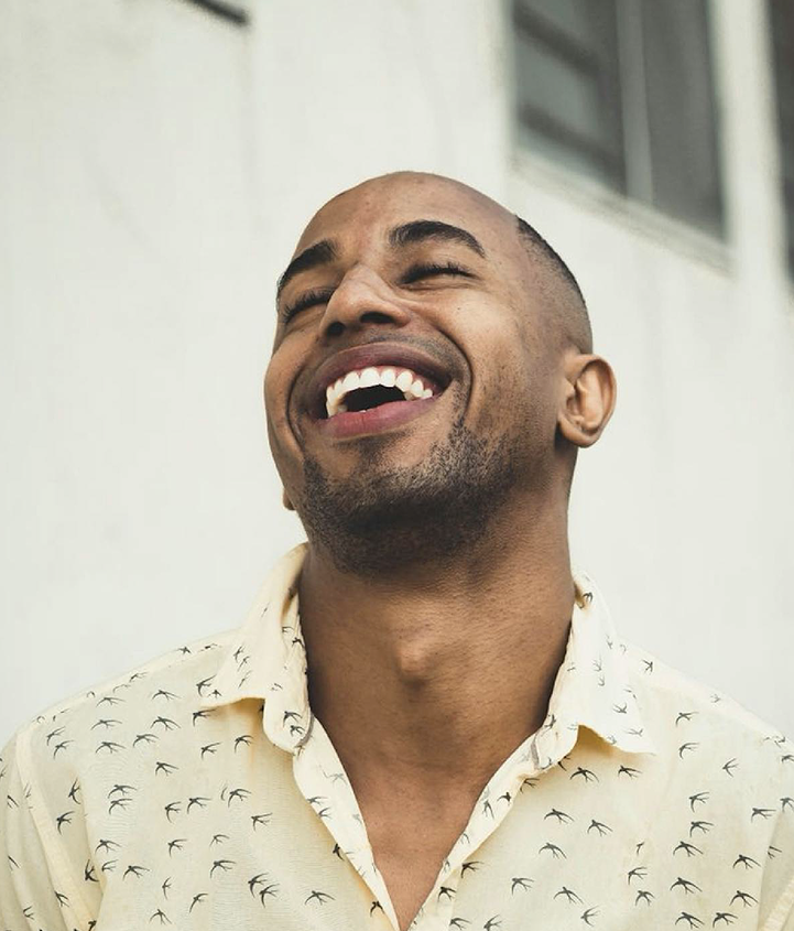 A person is standing outdoors in front of a light-colored building with large windows. They are wearing a light beige button-up shirt with a pattern of small black birds scattered across it. The background suggests a modern, urban setting with soft natural light.