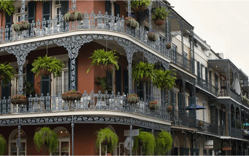 A multi-story brick building with ornate iron balconies, typical of New Orleans architecture, is adorned with numerous hanging plants and flower baskets. The balconies wrap around the corner of the building, featuring intricate railings and green shutters on the windows. Below, there are storefronts with signs, including one for a "Discount T-Shirt Center" and another indicating an ATM inside, set against a backdrop of a historic urban street.