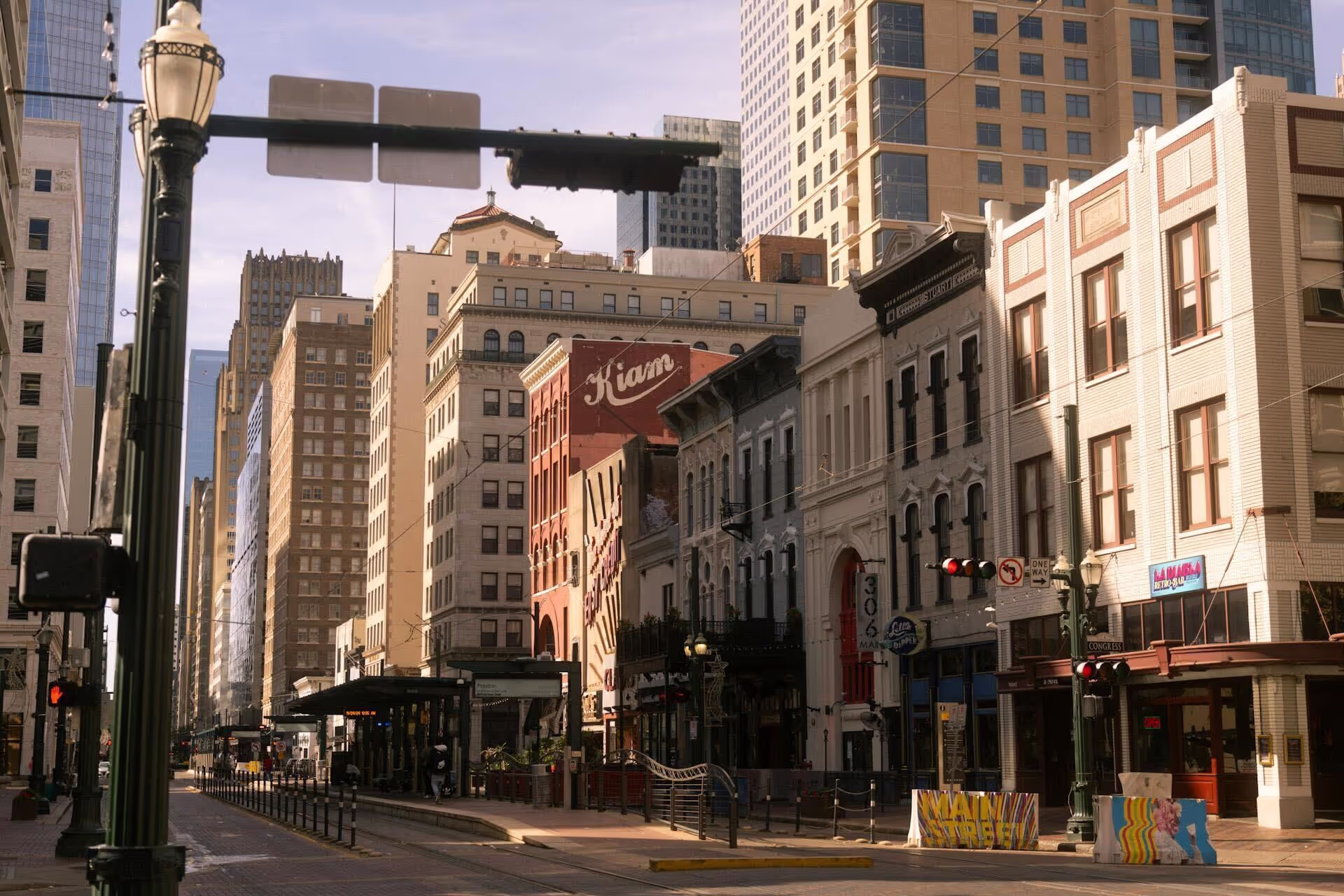 Urban street scene in downtown Houston, Texas, featuring historic buildings along Main Street, including the Kiam Building with its name painted on a red brick wall. A METRORail station and streetcar tracks run through the center of the street, with a mix of older and modern high-rise buildings in the background.