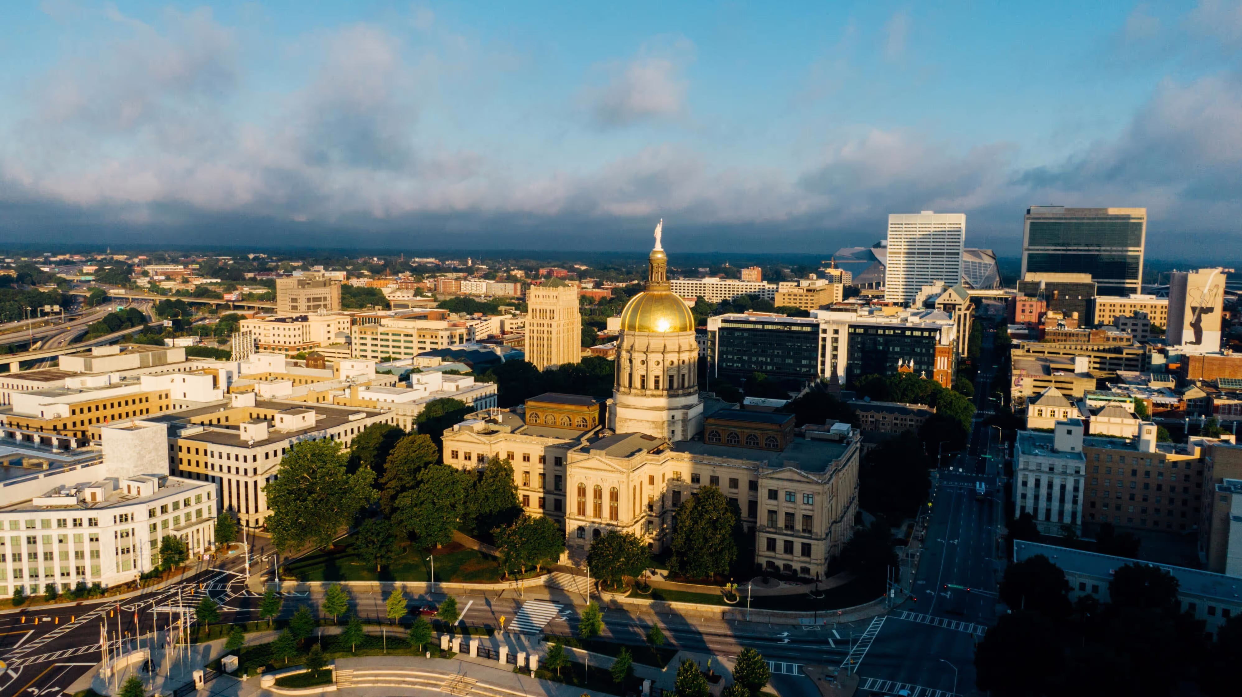 Aerial view of downtown Atlanta, Georgia, at sunrise, featuring the Georgia State Capitol with its prominent gold dome in the foreground. Modern office buildings and stadiums are visible in the background under a partly cloudy sky, with tree-lined streets and government buildings surrounding the capitol.