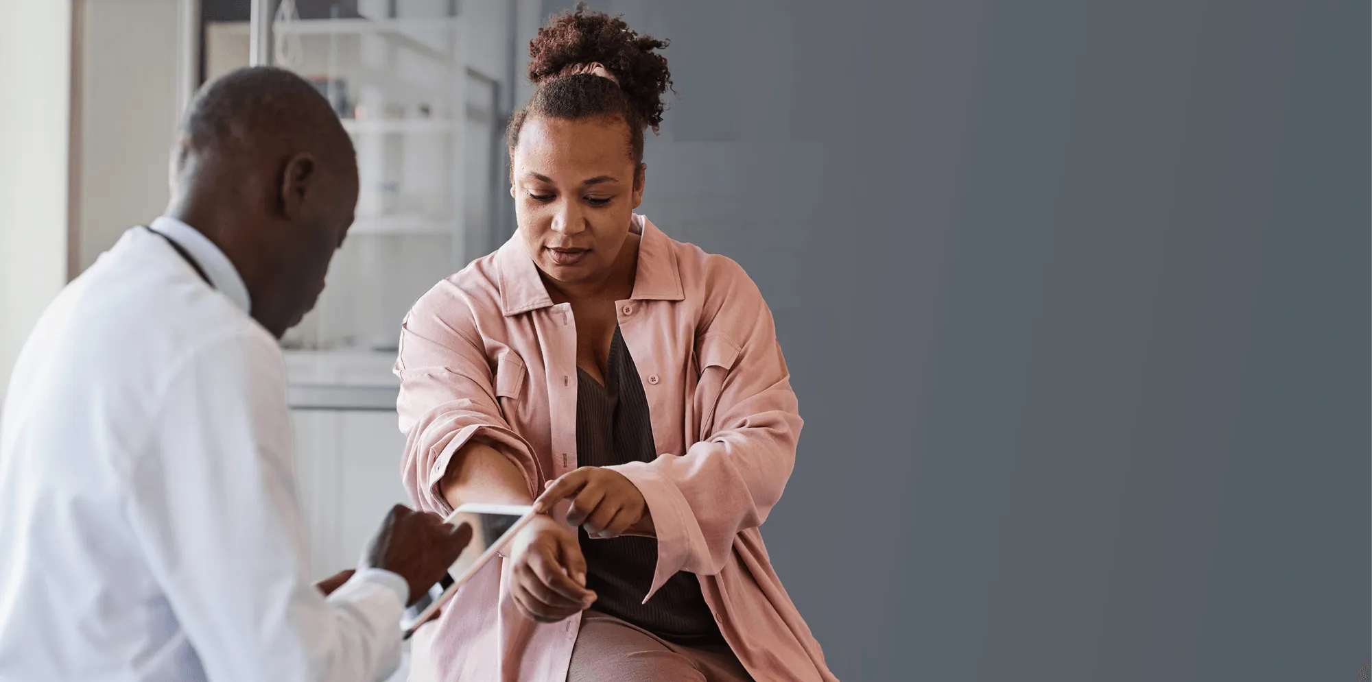 A female patient pointing to her arm while speaking with a male doctor using a tablet.