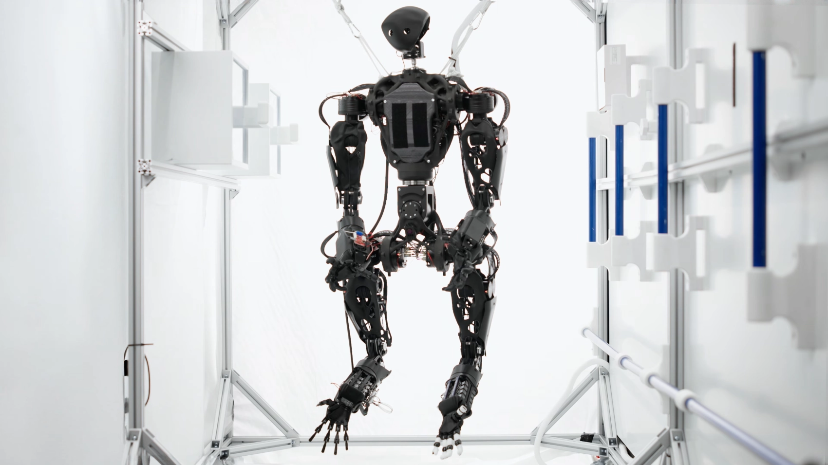 Černý humanoidní robot Helios s mechanickou konstrukcí, který visí na popruzích uprostřed bílé testovací laboratoře.