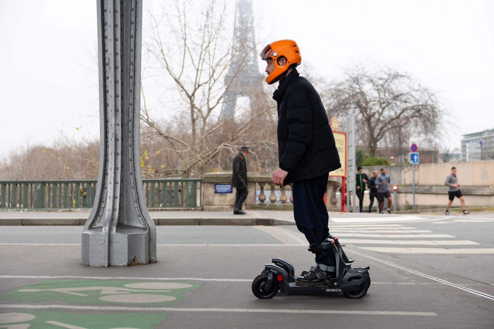 Muž v oranžové helmě jede na elektrických bruslích Skwheel Peak po městské cyklostezce s Eiffelovou věží v pozadí.