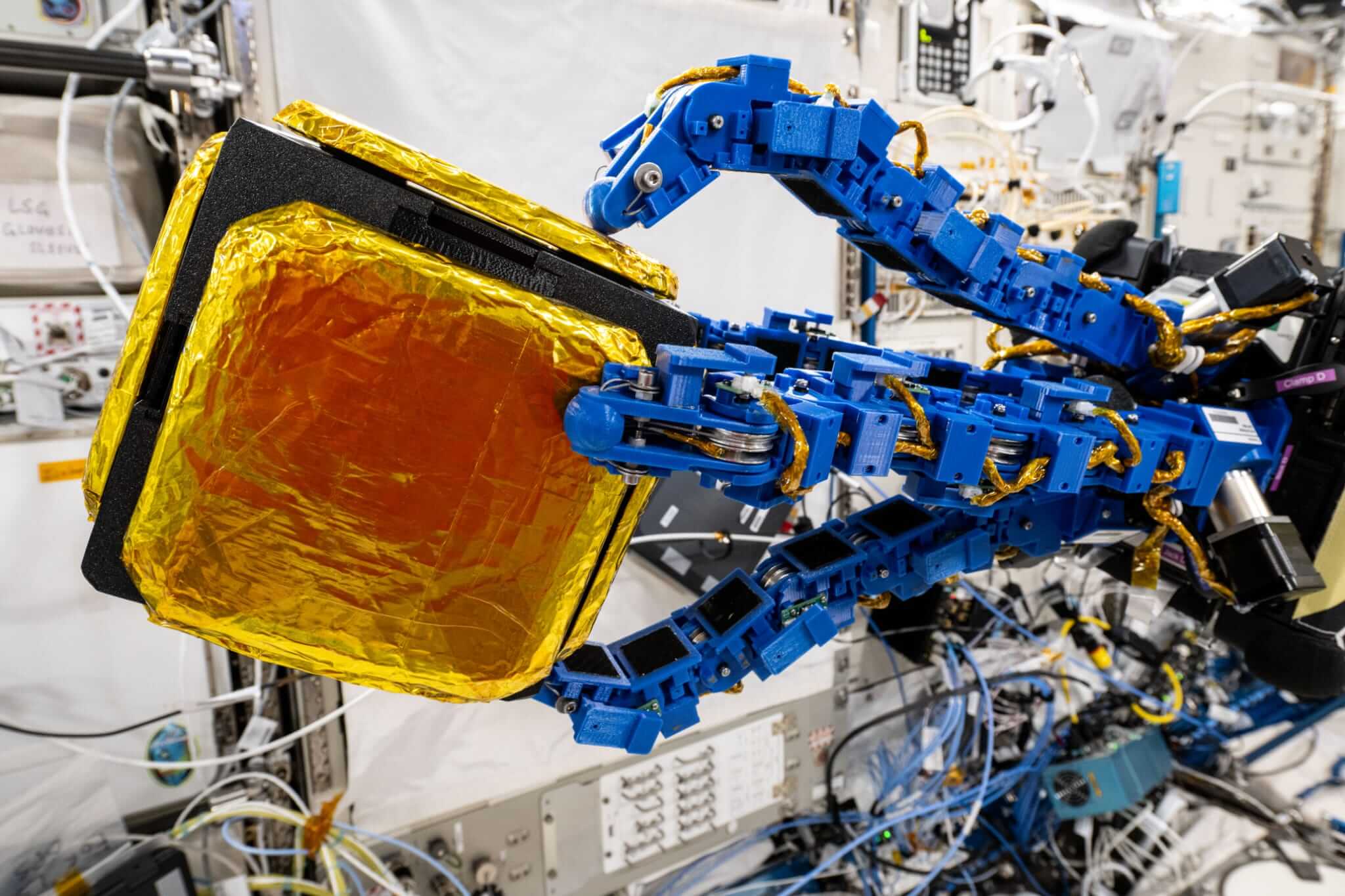 Modrá chapadlovitá ramena s přísavkami podobnými těm, jaké má gekon, připevněná k robotickému letounu Astrobee, se natahují a uchopují „záchytný krychlový modul“ uvnitř laboratorního modulu Kibo na Mezinárodní vesmírné stanici. 