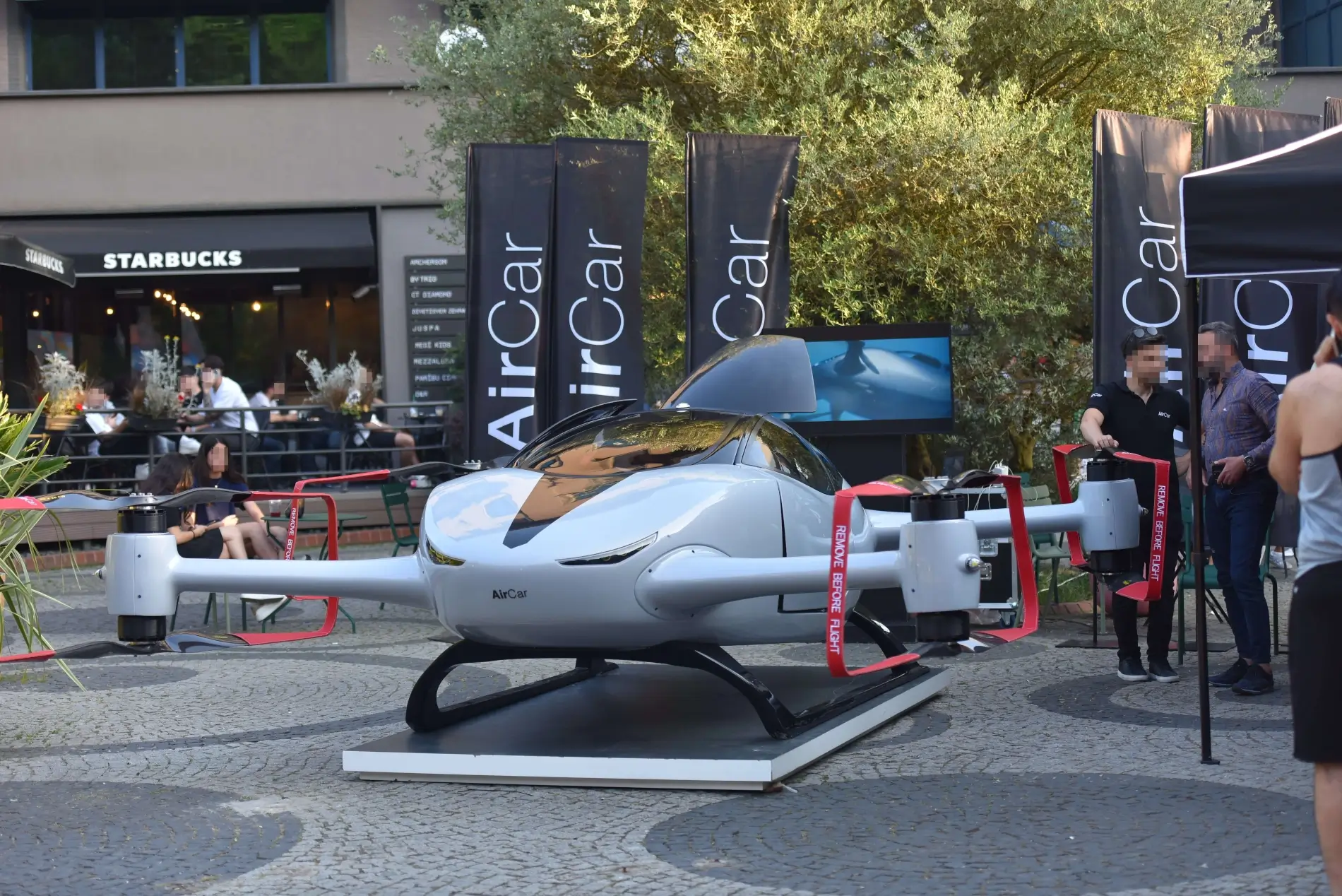 Elektrický osobní dron AirCar vystavený na veřejném prostranství před kavárnou Starbucks s rozmazanými obličeji osob v pozadí.